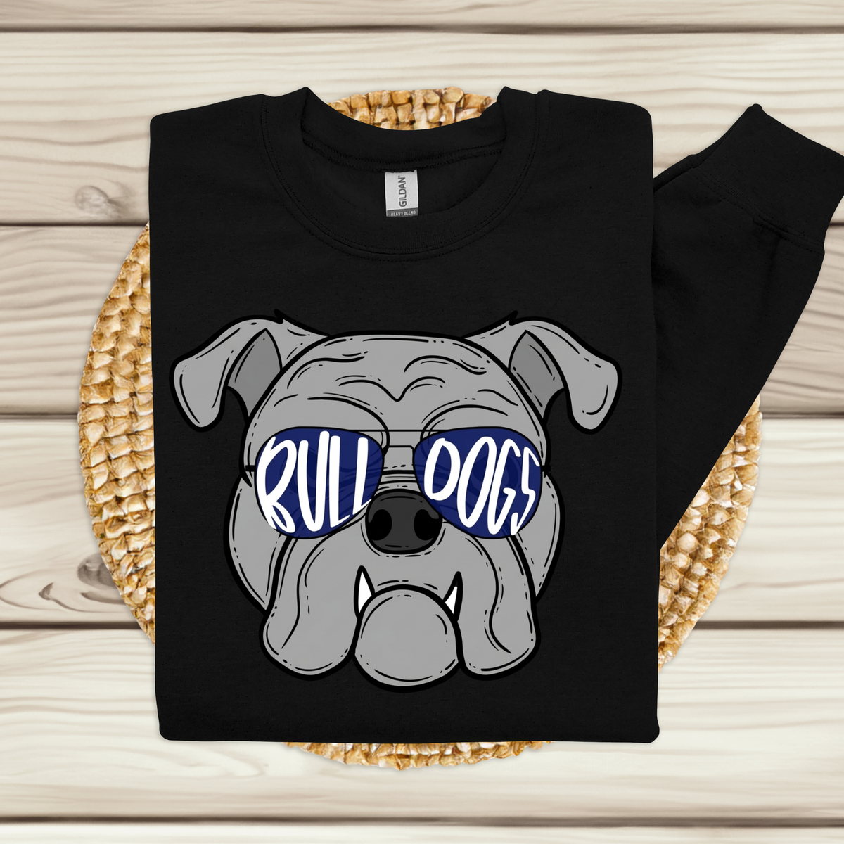 A black long-sleeve sweatshirt laid flat on a woven round mat over a light wood plank surface. The sweatshirt features a large graphic of a gray bulldog wearing blue sunglasses with the words 'RULL DOGS' in white.