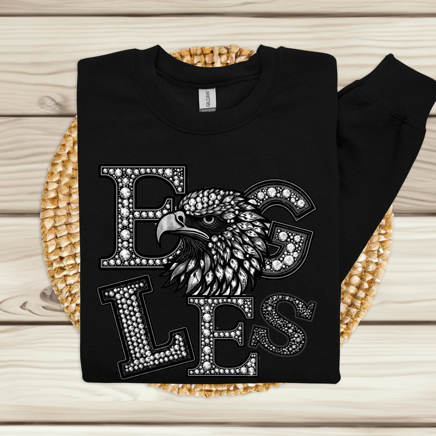 A black long-sleeve crewneck sweatshirt laid flat on a woven round mat over a light wood plank background. The front features a large graphic with the word 'EGLES' in decorative stone-like letters and an eagle head integrated into the design.