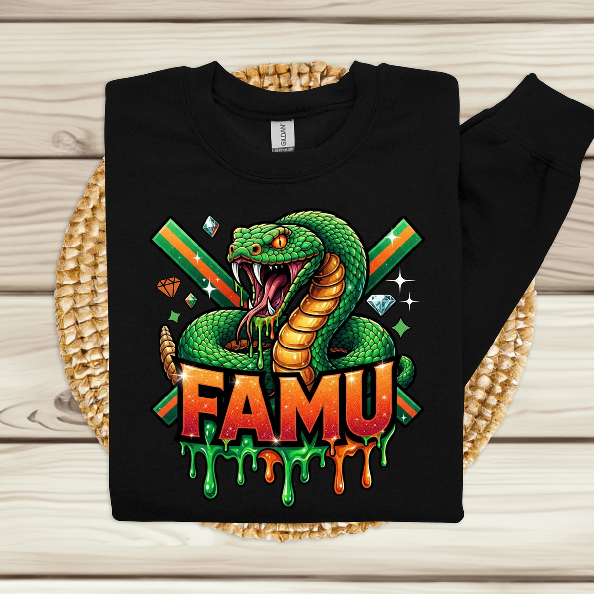 A black long-sleeve sweatshirt laid flat on a woven round mat over a light wood plank background. The sweatshirt features a large colorful graphic of a green snake with its mouth open, crossed green and orange diagonal stripes behind it, gem icons, and the word 'FAMU' in dripping orange and green letters.