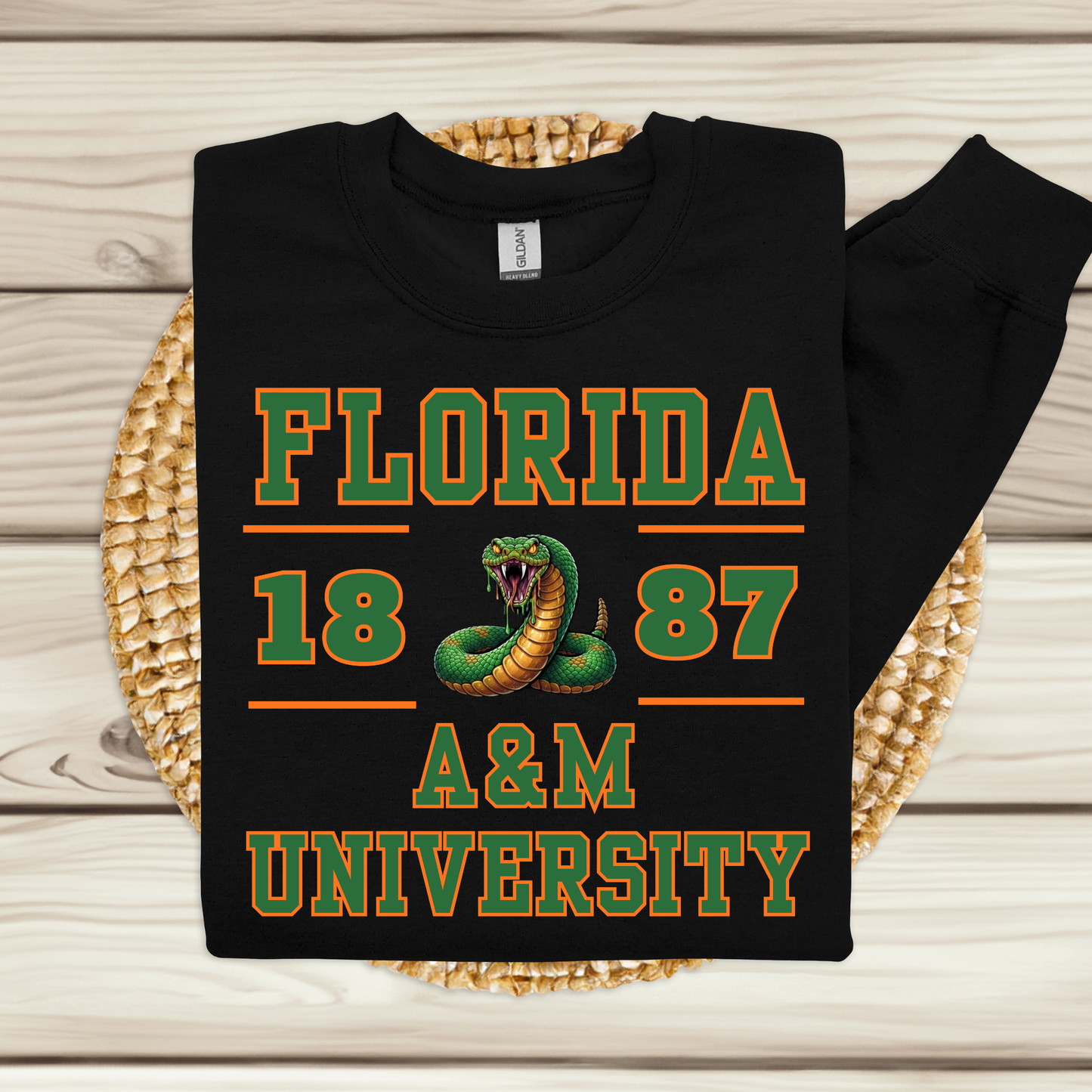 A black long-sleeve sweatshirt with a Florida 1887 A&M University graphic, displayed flat on a woven round mat over a light wood plank background.