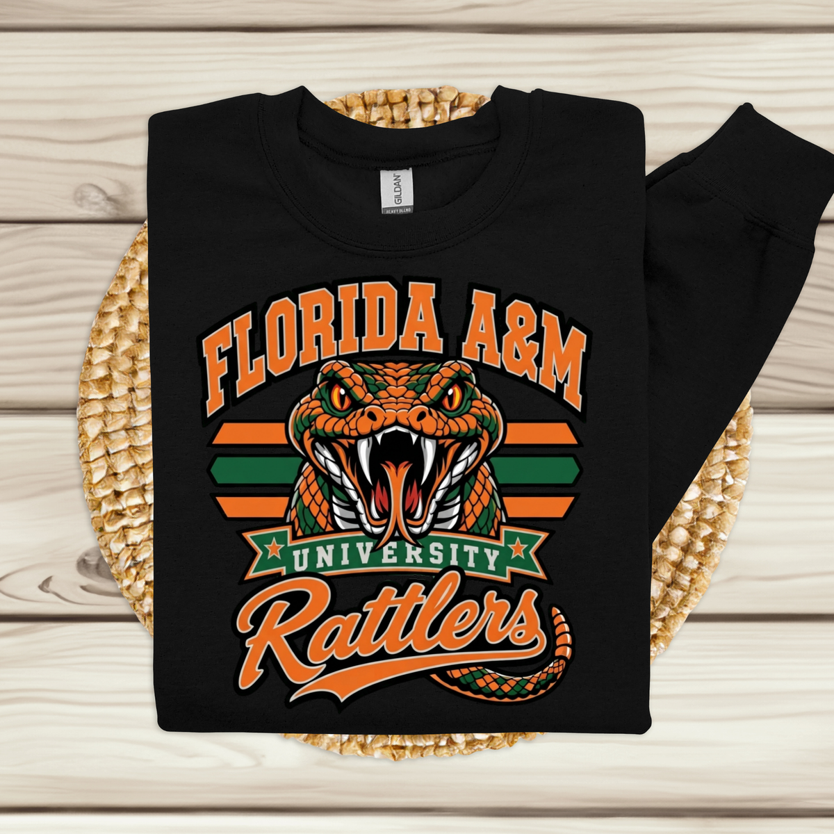 A black long-sleeve sweatshirt with a Florida A&M University Rattlers graphic, displayed flat on a woven straw mat over a light wood plank background.