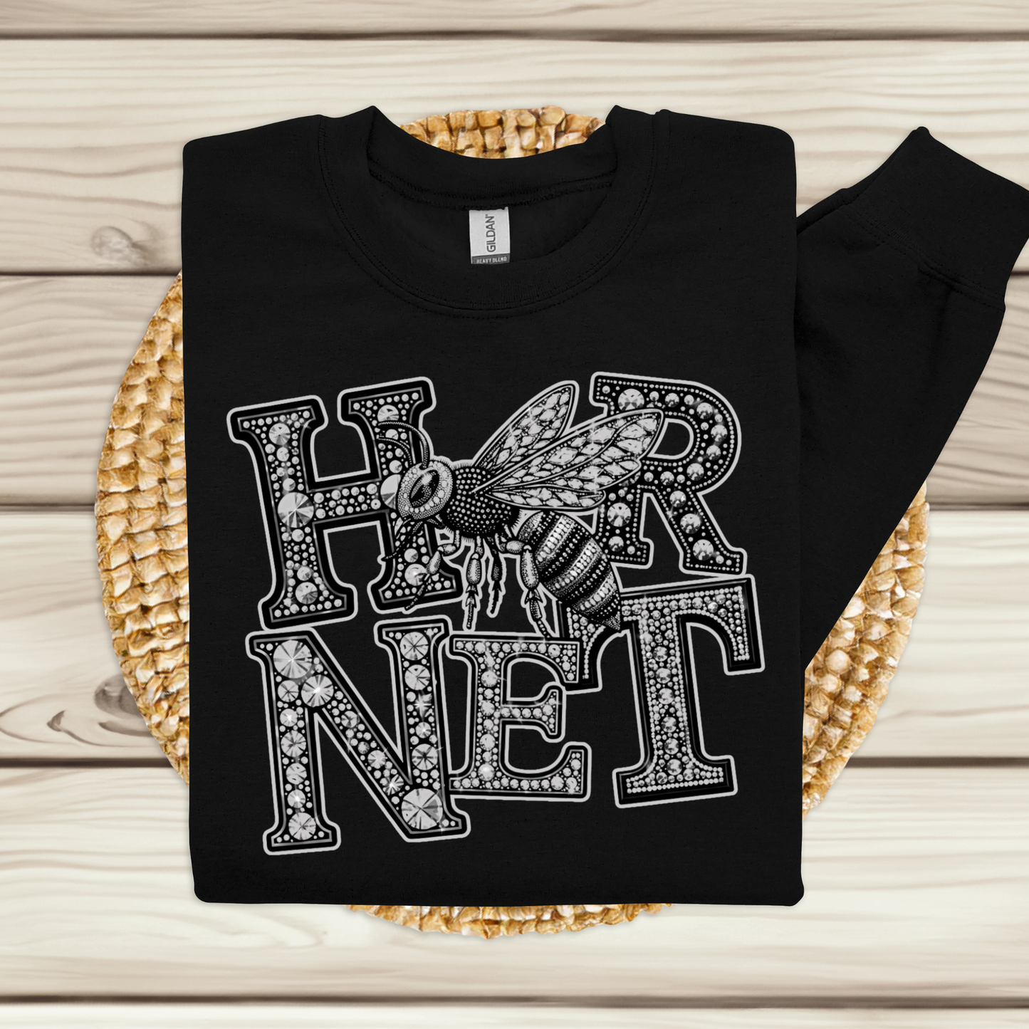 A black long-sleeve crewneck sweatshirt laid flat on a woven straw mat over light wood planks. The front has a large white and gray graphic spelling 'HORNET' with a detailed bee illustration integrated into the lettering.