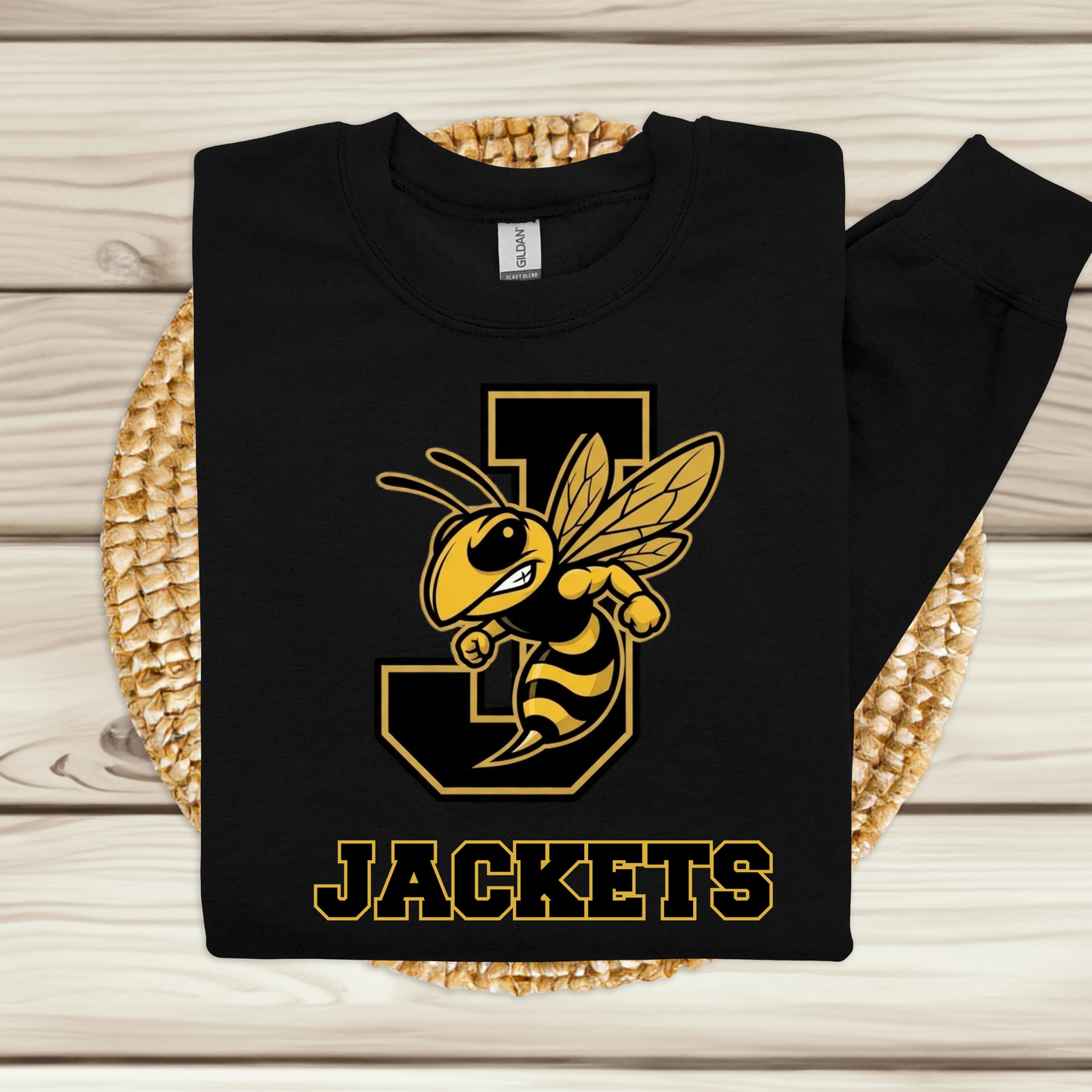 A black long-sleeve sweatshirt with a yellow and black 'Jackets' bee mascot graphic, laid flat on a woven placemat over a light wood background.