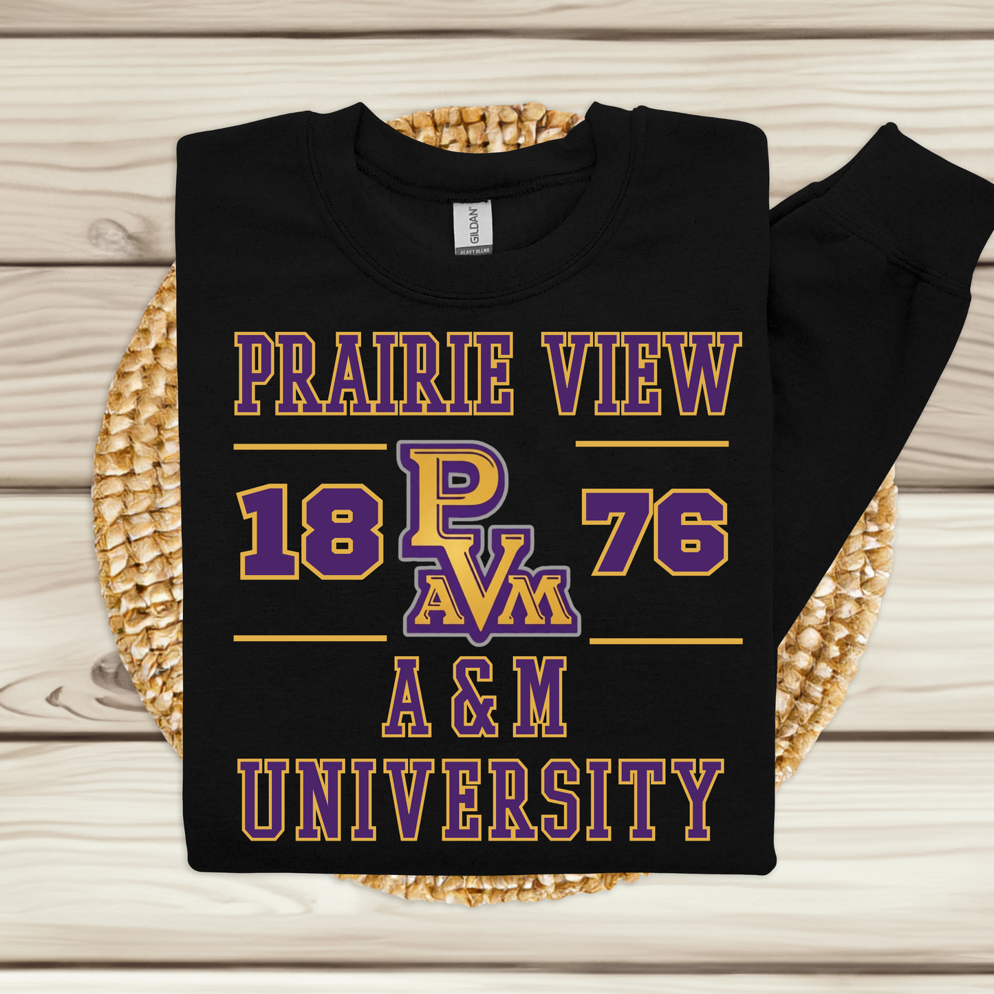 A black long-sleeve t-shirt with a purple and gold Prairie View A&M University graphic, displayed flat on a woven placemat over a light wood plank background.
