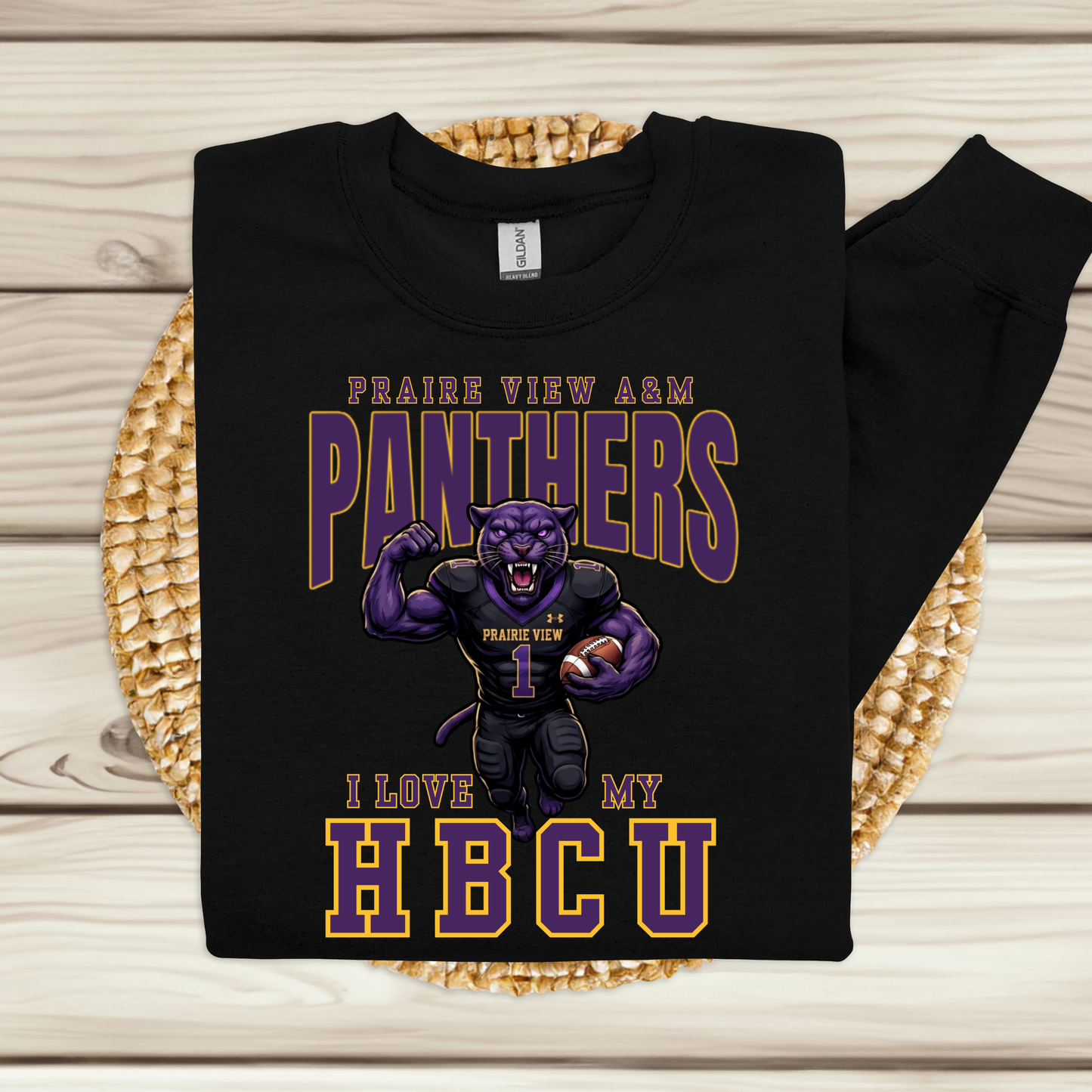 A black sweatshirt laid flat on a woven round mat over a light wood plank background. The sweatshirt features a large graphic with the words 'PRAIRIE VIEW A&M PANTHERS I LOVE MY HBCU' and an illustrated purple panther mascot wearing a football uniform with the number 1.