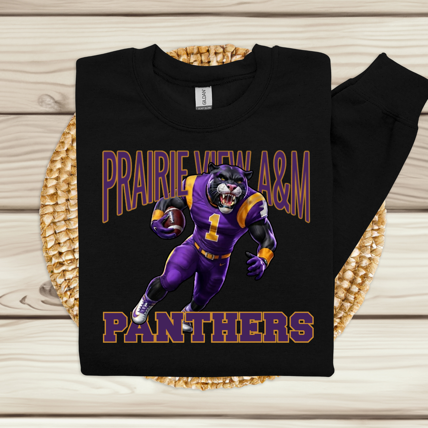 A black long-sleeve sweatshirt with a purple and gold 'Prairie View A&M Panthers' football graphic, displayed flat on a woven placemat over a light wood background.