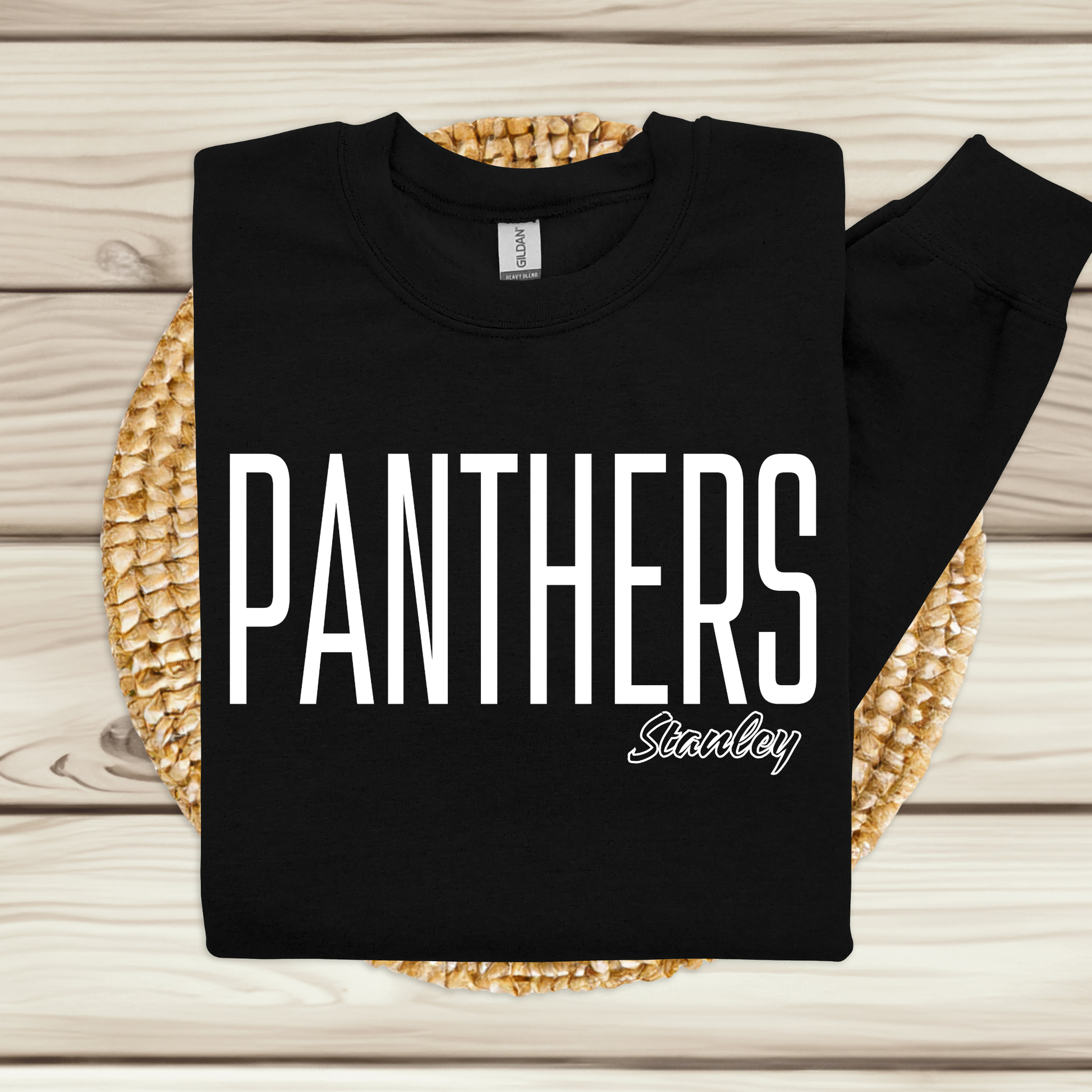 A black long-sleeve sweatshirt with 'PANTHERS Stanboy' printed in white across the front, laid flat on a woven round mat over a light wood plank background.
