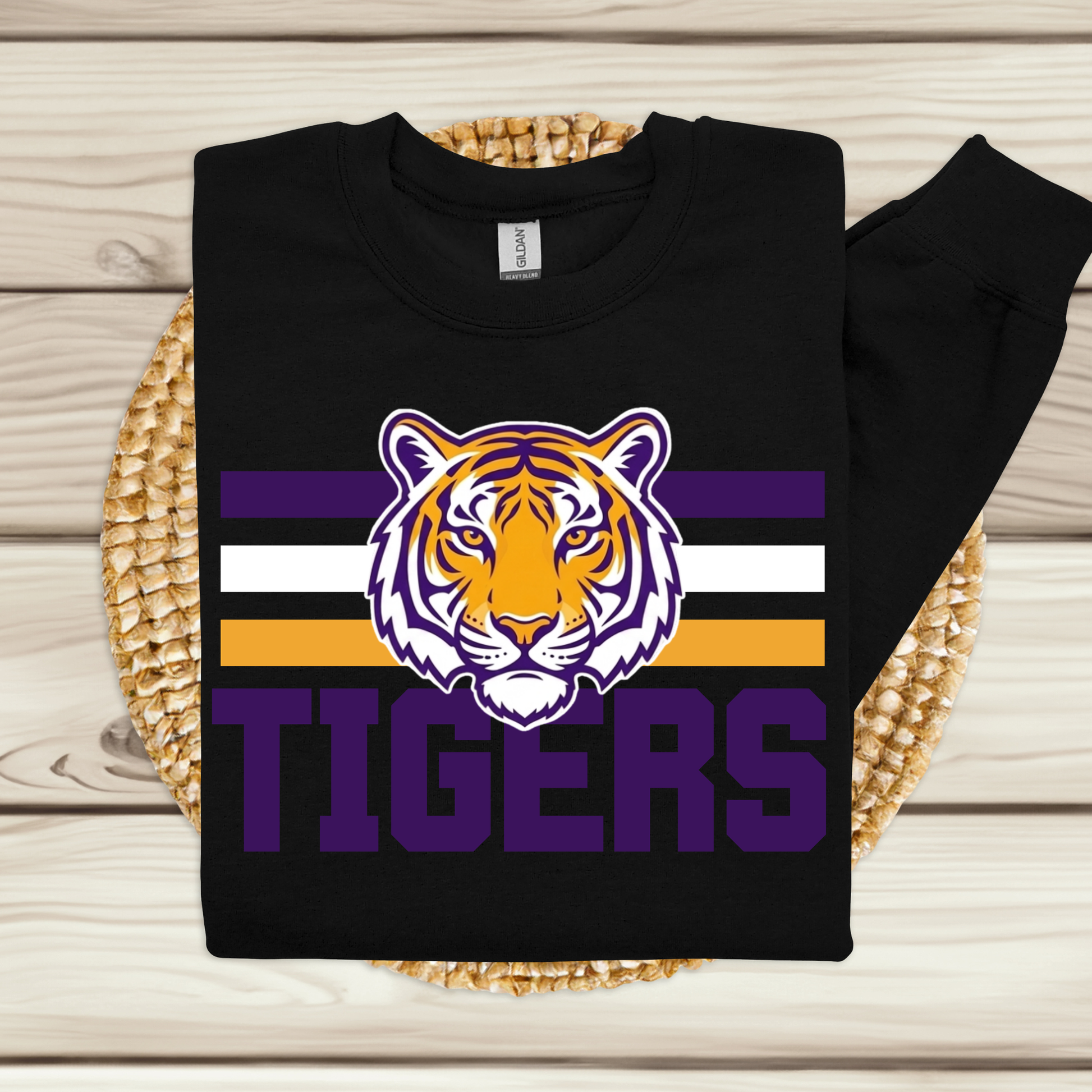 A black long-sleeve sweatshirt laid flat on a woven round mat over a light wood plank surface. The sweatshirt features a large tiger head graphic above the word "TIGERS" in purple, with purple, white, and gold horizontal stripes behind the graphic.