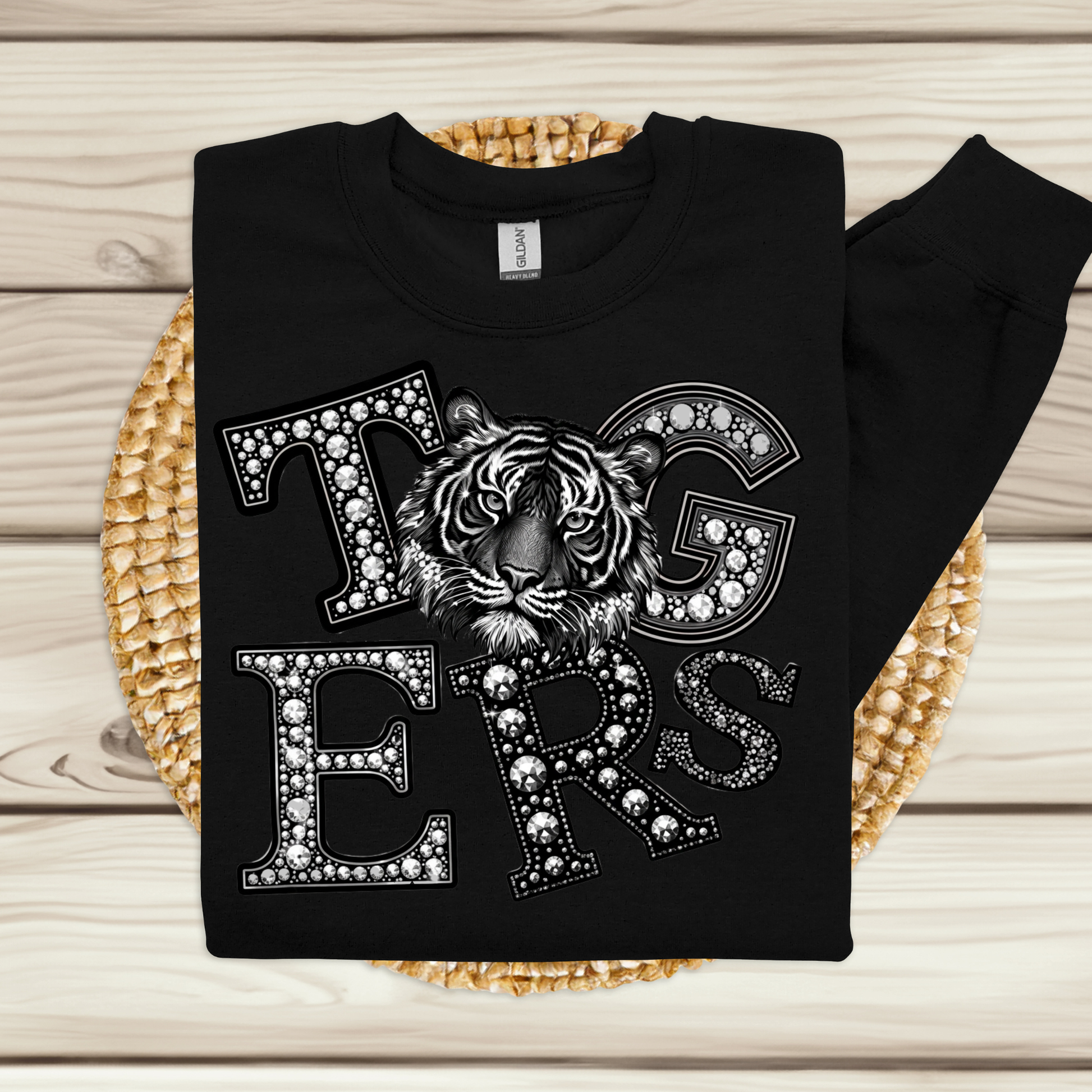 A black long-sleeve sweatshirt laid flat on a woven round mat over a light wood plank surface. The front has a large graphic reading "Tigers" in outlined letters filled with rhinestone-like dots, with a detailed tiger face across the letters.