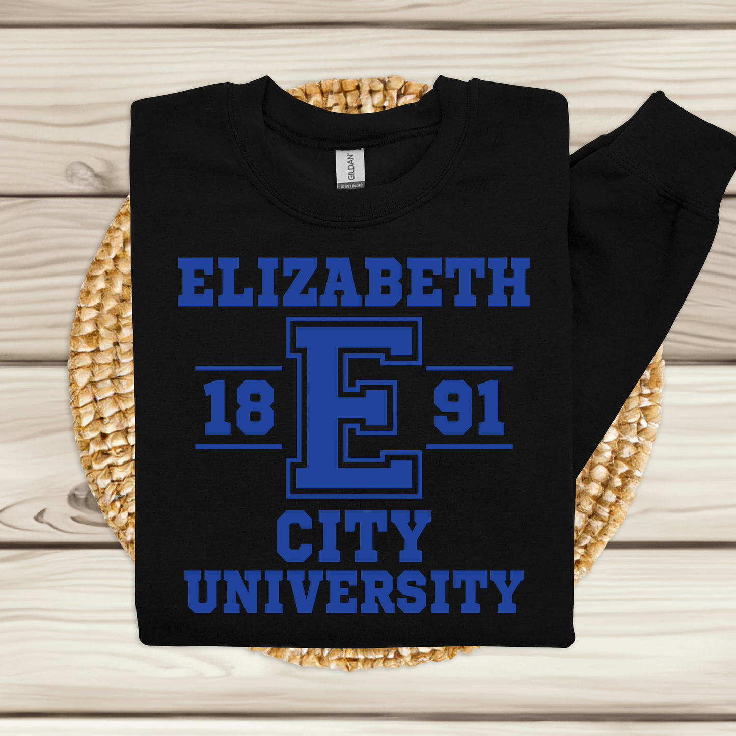 A black long-sleeve sweatshirt laid flat on a woven round mat over a light wood plank surface. The sweatshirt has a large blue graphic print reading 'ELIZABETH E 1891 CITY UNIVERSITY' with a large block letter E in the center.