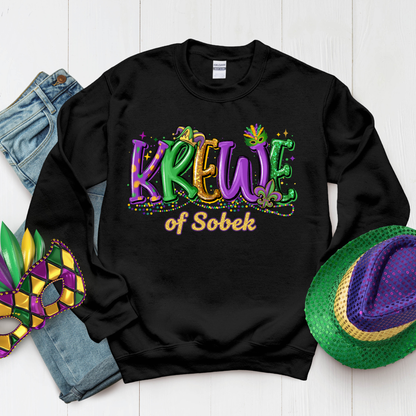 Black sweatshirt with 'Krewe of Sobek' text, surrounded by Mardi Gras accessories on a white background