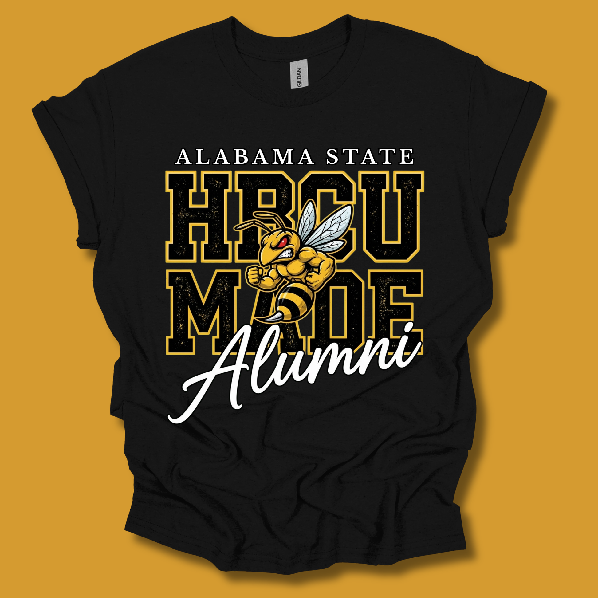 A black graphic t-shirt featuring the text 'ALABAMA STATE HBCU MADE Alumni' with a cartoon bee mascot, displayed against a solid mustard yellow background.