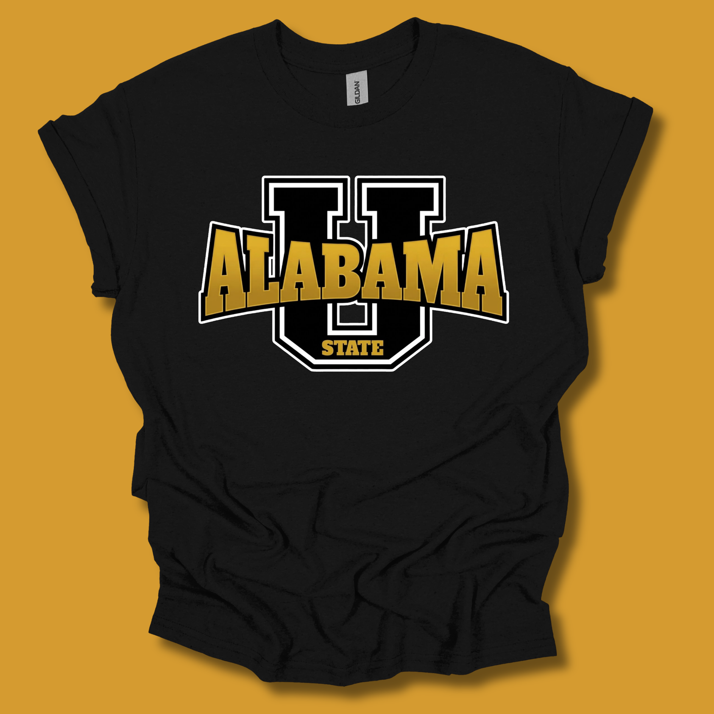 A black short-sleeve t-shirt with a graphic of a large black and white letter 'U' outlined with 'ALABAMA STATE' in gold and white. The shirt is displayed flat against a solid mustard yellow background.