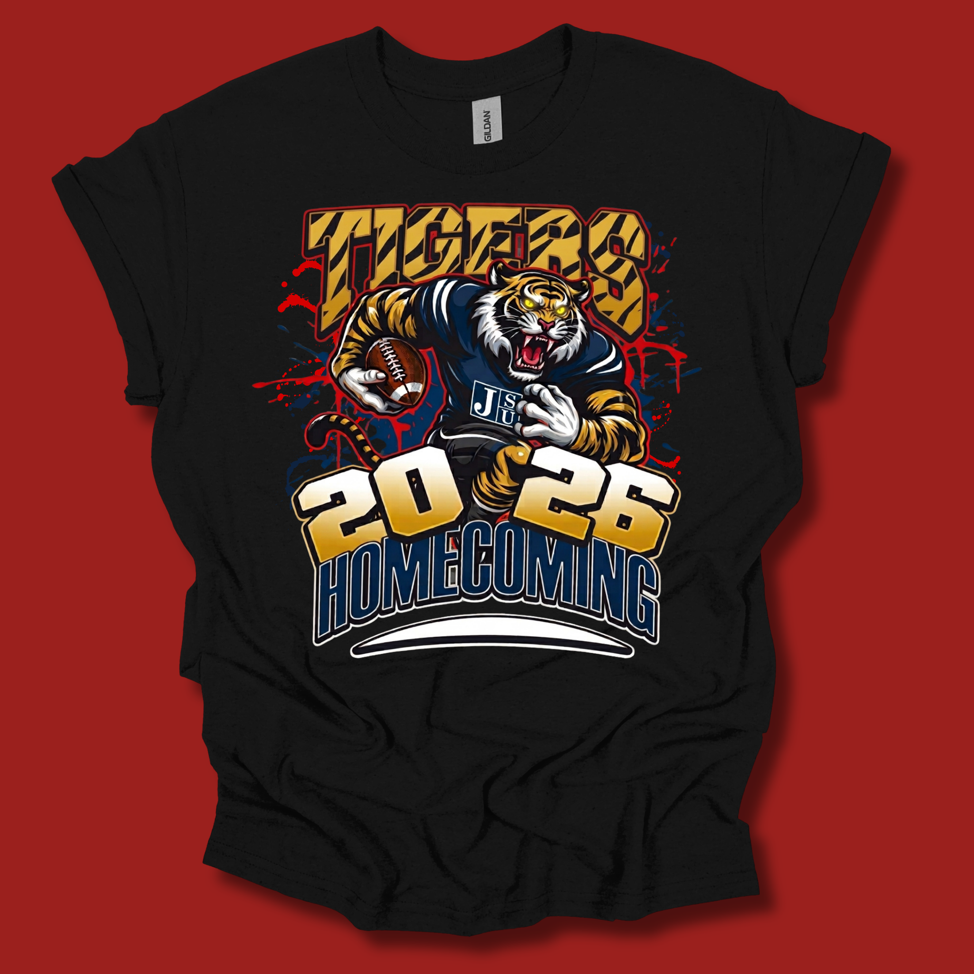 A black short-sleeve graphic t-shirt featuring a tiger mascot holding a football, with 'TIGERS' and '2026 HOMECOMING' printed in bold yellow, blue, and red lettering. The t-shirt is displayed flat against a solid red background.
