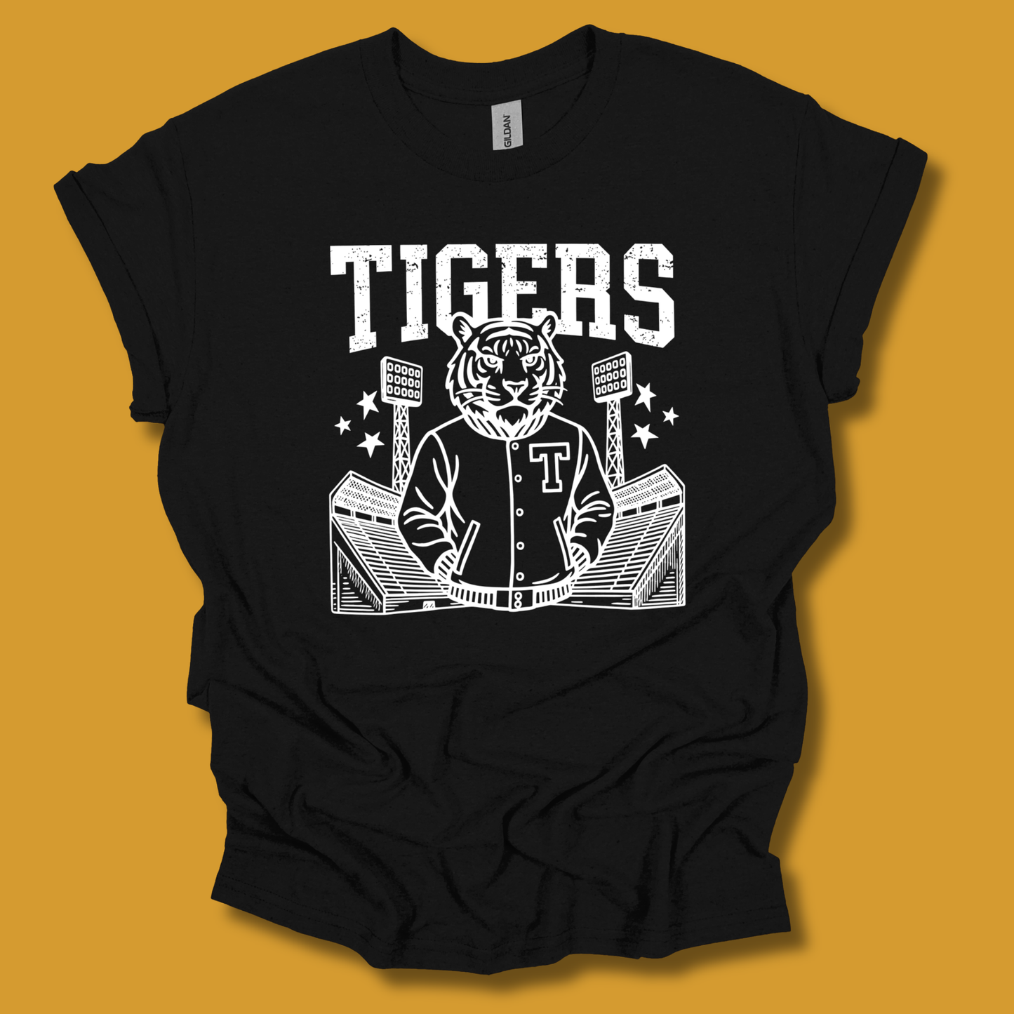 A black short-sleeve t-shirt with a white graphic print of a tiger mascot in a varsity jacket at a stadium, with the word 'TIGERS' above, shown against a solid mustard yellow background.