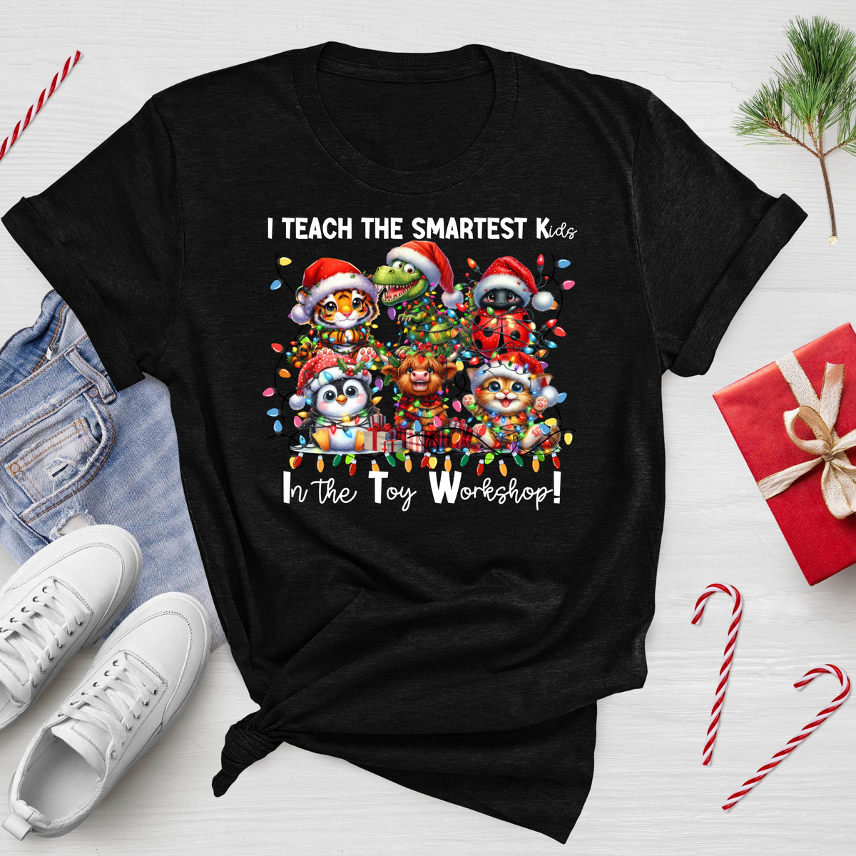 A black graphic t-shirt featuring a colorful illustration of Christmas-themed toy animals wearing Santa hats, with the text 'I TEACH THE SMARTEST Kids In The Toy Workshop!'. The t-shirt is styled in a flat-lay arrangement on a white wooden background, surrounded by blue jeans, white sneakers, a red gift box, candy canes, and a small pine branch.