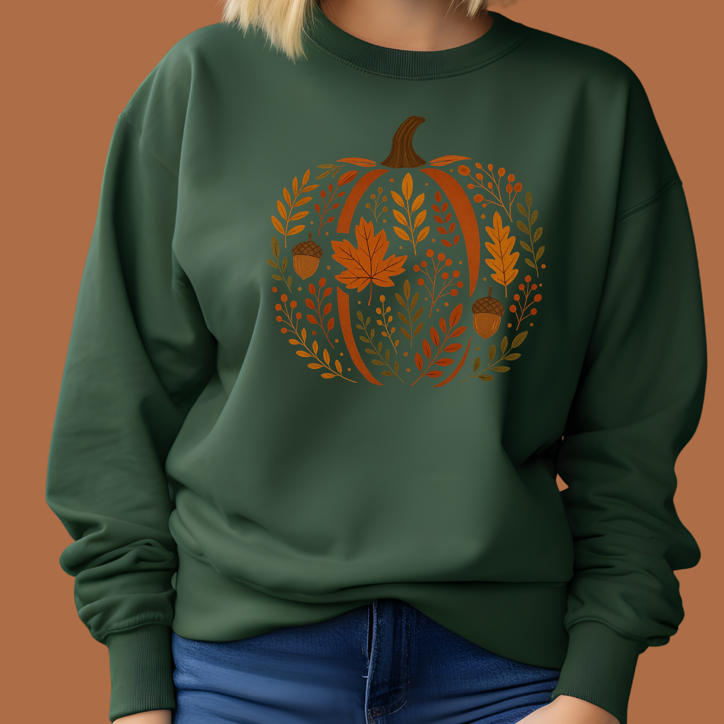A person wearing a dark green crewneck sweatshirt with a large autumn-themed pumpkin graphic in orange, brown, and gold tones, paired with blue jeans against a solid warm brown background.