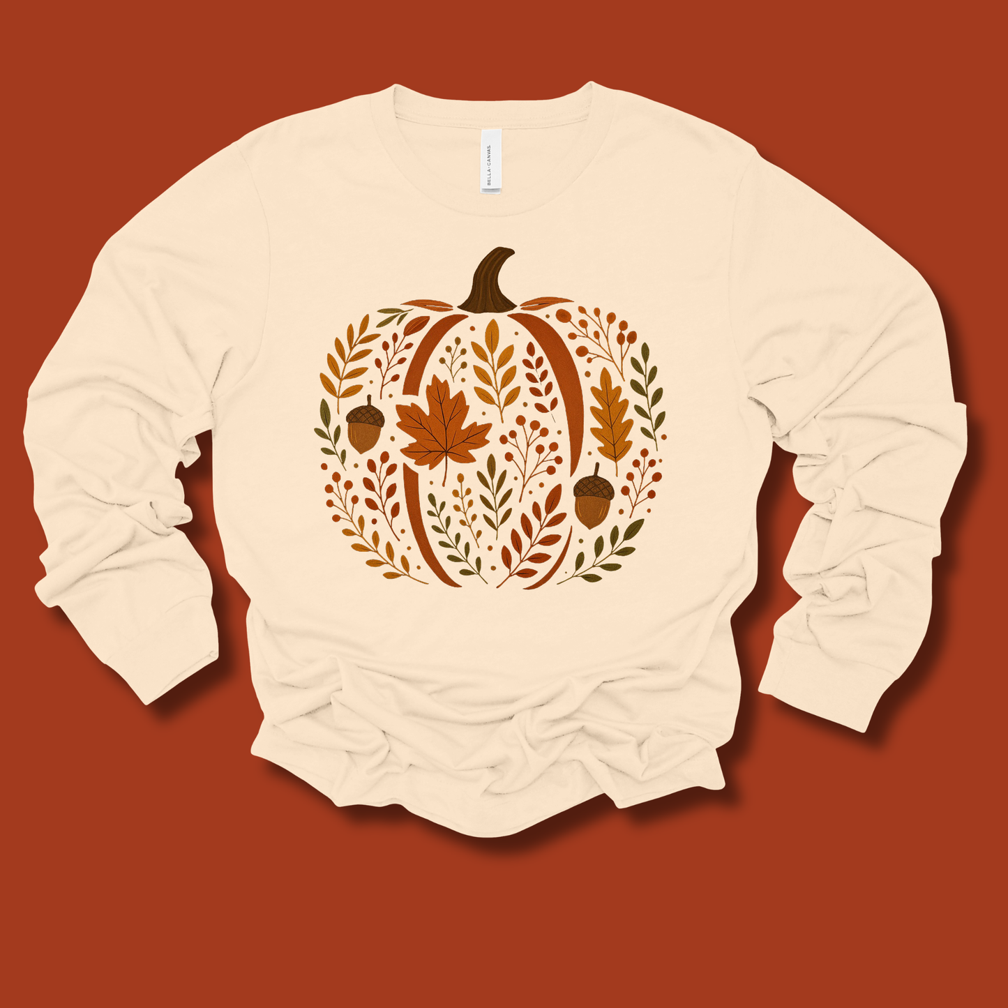 A cream-colored long-sleeve t-shirt with a fall-themed pumpkin graphic made of leaves, acorns, and maple leaves, displayed flat against a solid terracotta background.