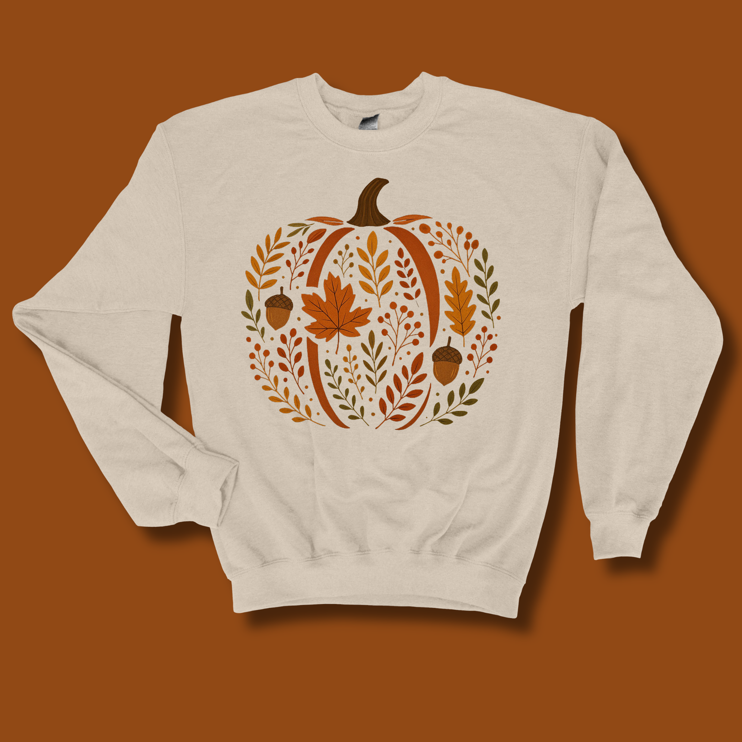 An off-white crewneck sweatshirt featuring a large autumn-themed pumpkin graphic on the chest, laid flat against a solid brown background.