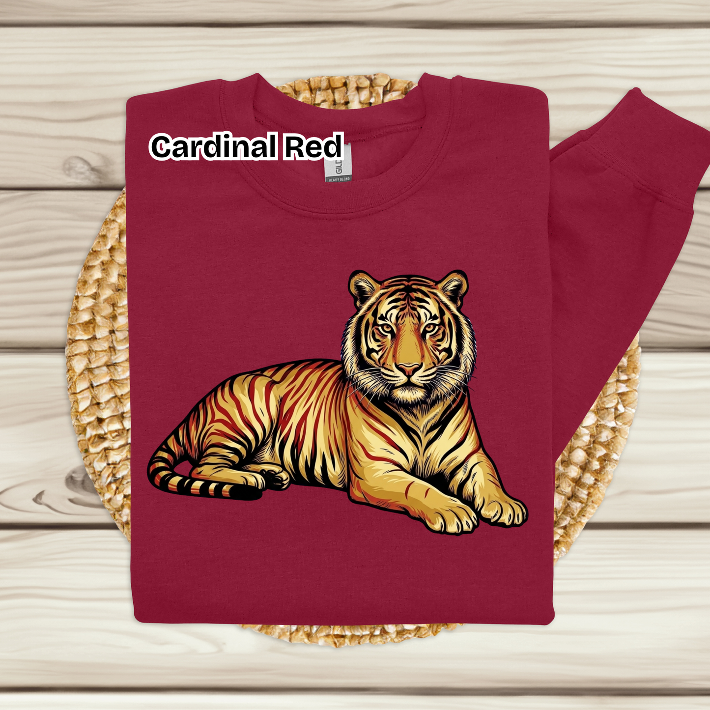 A folded crewneck sweatshirt in cardinal red, featuring a graphic print of a resting tiger on the front. The sweatshirt is displayed on a round woven straw mat over a light-colored wooden plank background.