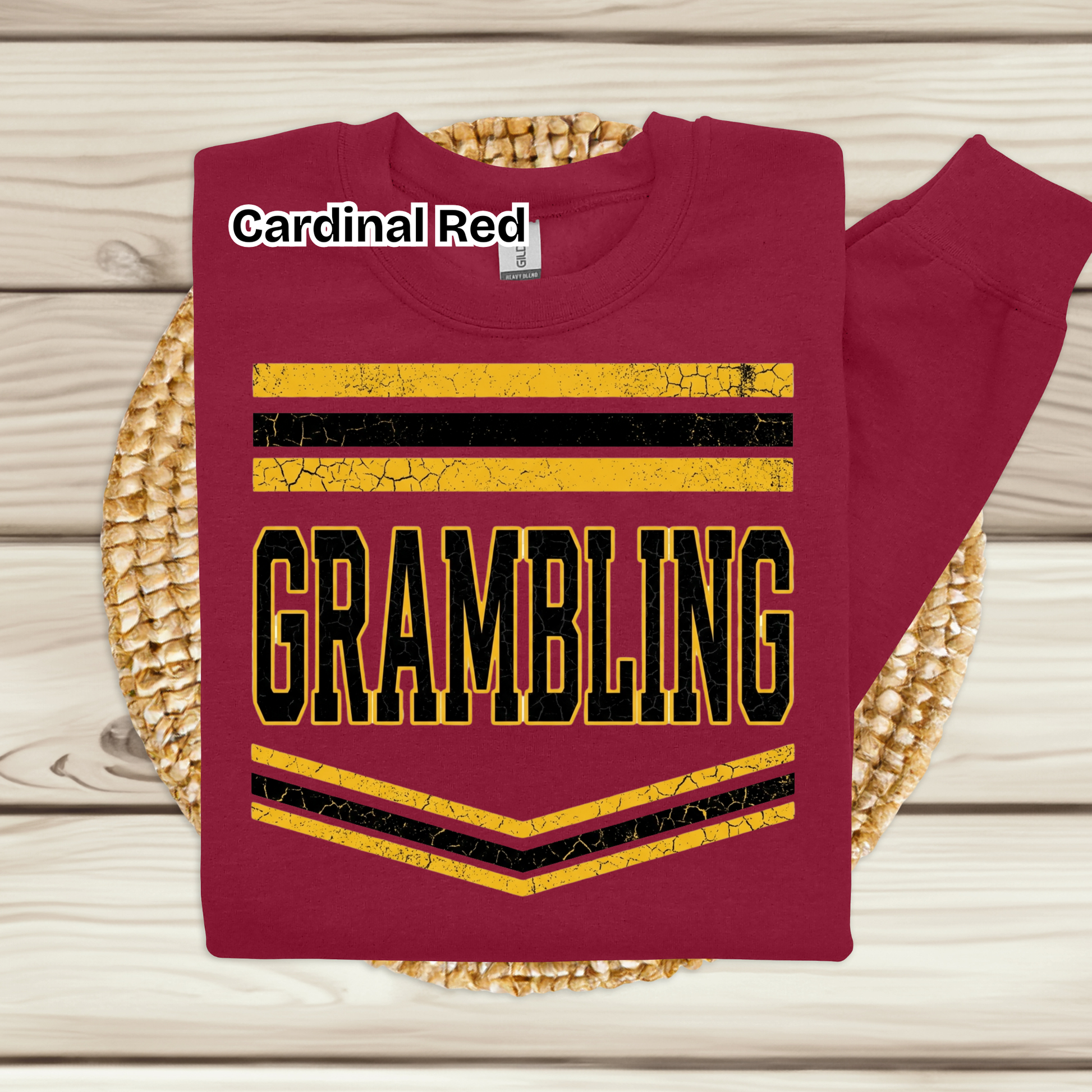 A folded crewneck sweatshirt in a deep cardinal red color, featuring a graphic design with yellow and black stripes and the word 'GRAMBLING' in bold block letters. The sweatshirt is displayed on a round woven placemat over a light wood plank background, with a text label overlay reading 'Cardinal Red'.
