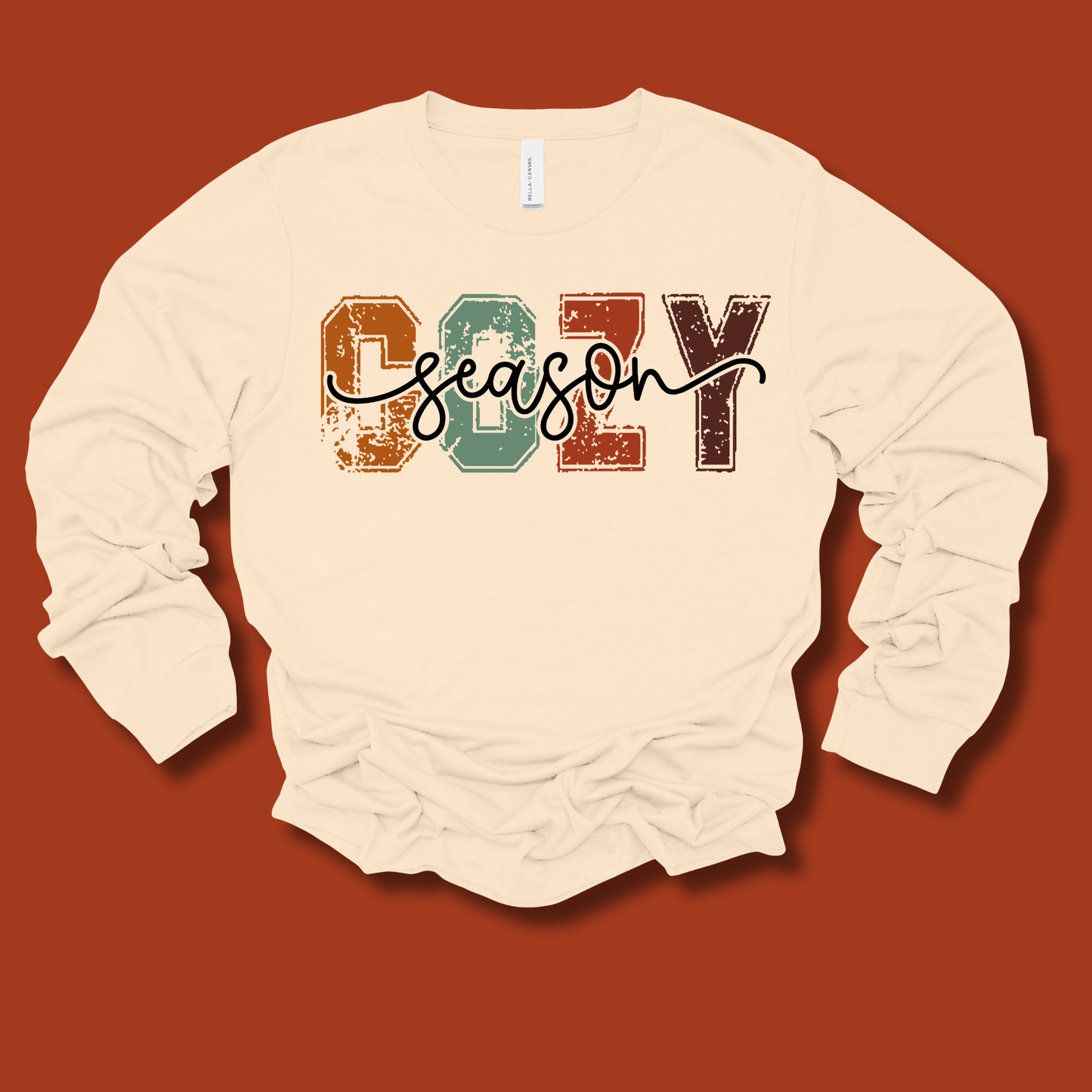 An off-white long-sleeve t-shirt with a distressed graphic reading 'COZY season' across the chest, laid flat against a solid terracotta background.
