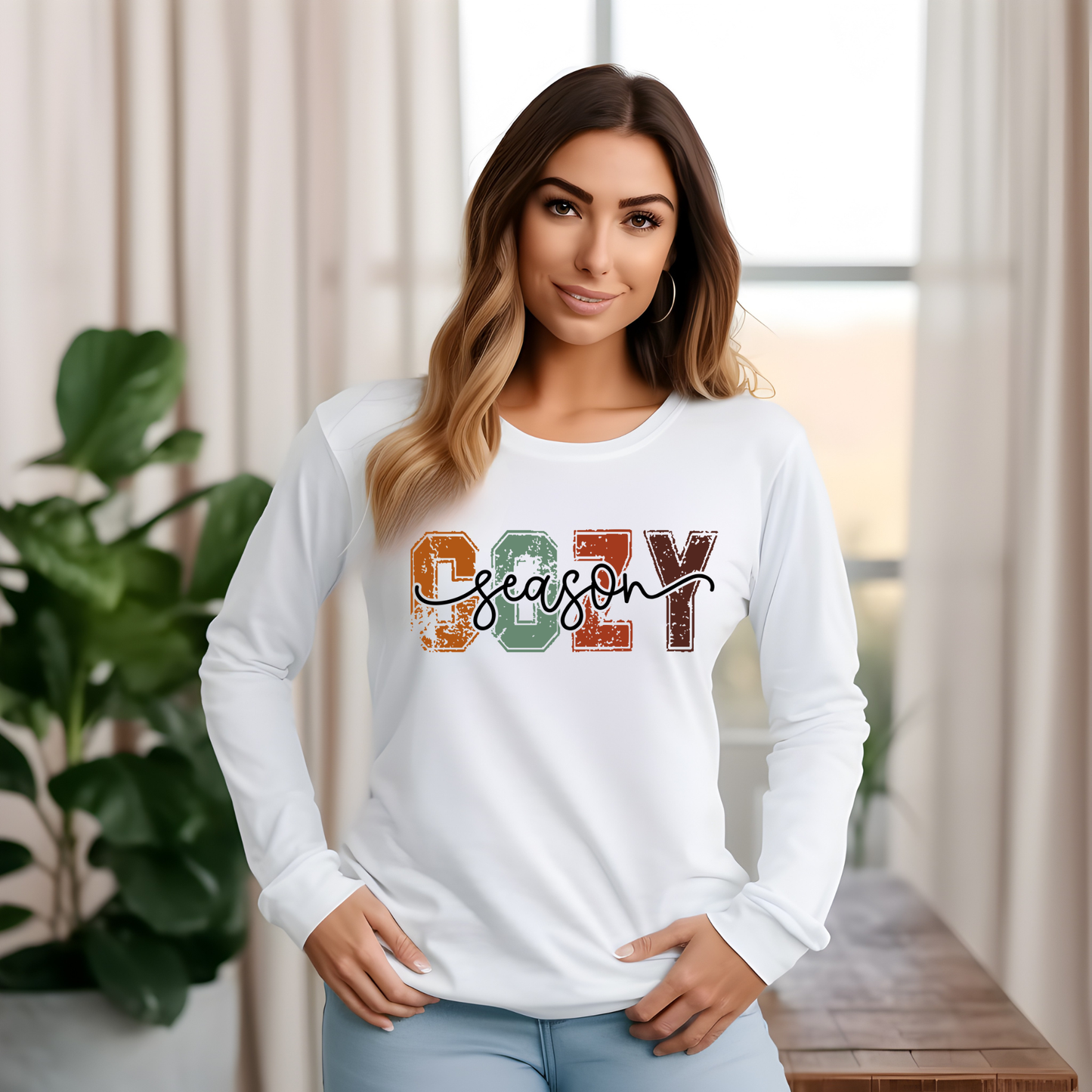 A woman wearing a white long-sleeve t-shirt with a colorful distressed graphic that reads 'COZY season' in autumn colors, standing in a brightly lit room with curtains and a plant.