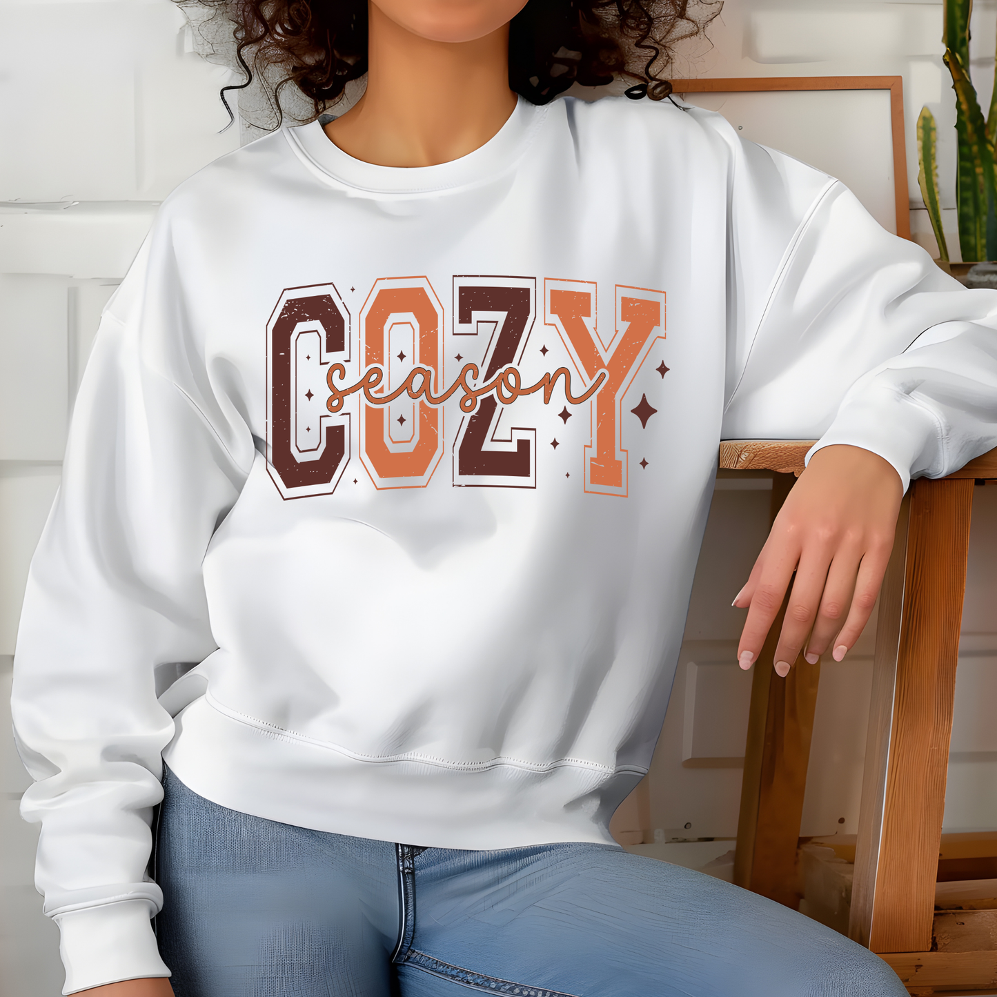 Person wearing a white sweatshirt with 'COZY season' text, sitting on a wooden stool.  Cozy Season Sweatshirt: Fall Vibe Retro Block Letter Crewneck Sweater