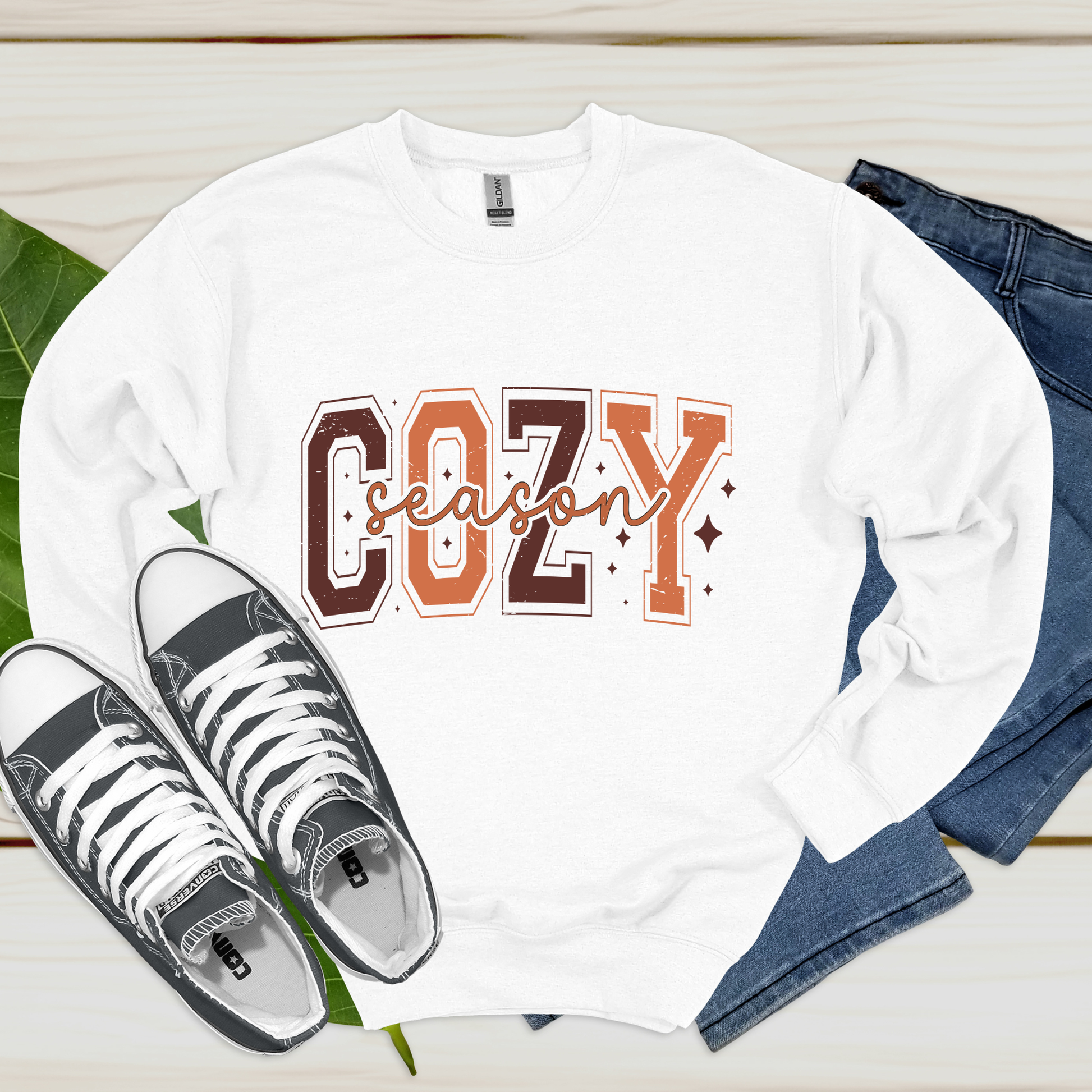 White long-sleeve shirt with 'Cozy Season' text, black high-top sneakers, and blue jeans on a wooden surface.  Cozy Season Sweatshirt: Fall Vibe Retro Block Letter Crewneck Sweater