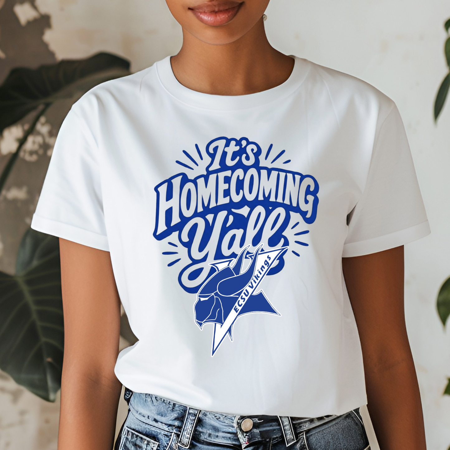 A woman wearing a white short-sleeve graphic t-shirt with a blue 'It's Homecoming Y'all' design featuring a rose and a ribbon that reads 'GO VILLAINS'.