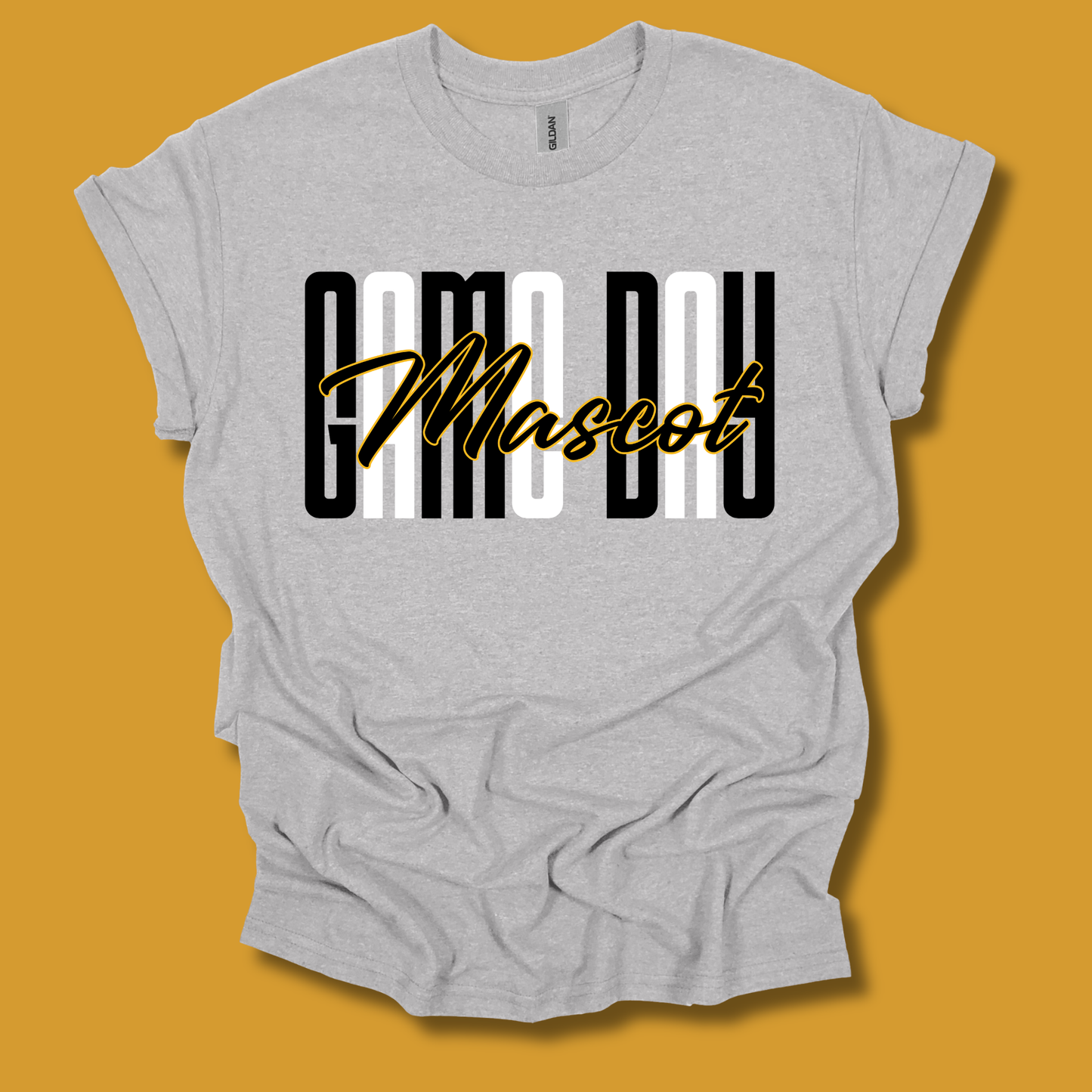 A light heather gray t-shirt with a graphic print reading 'CAMO DAY Mascot' in black, white, and gold, displayed against a solid mustard yellow background.