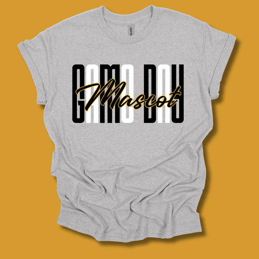 A light heather gray t-shirt with a graphic print reading 'CAMO DAY Mascot' in black, white, and gold, displayed against a solid mustard yellow background.