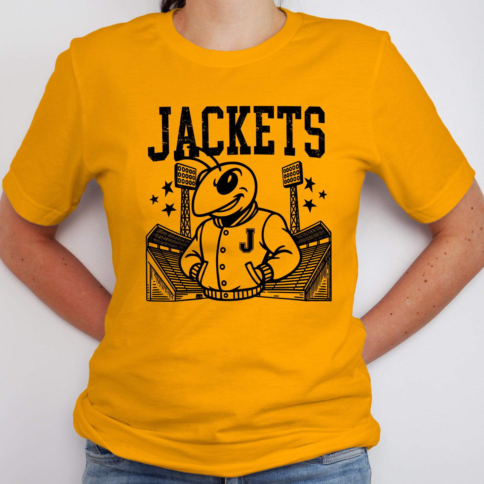 A person wearing a bright yellow short-sleeve t-shirt with a black graphic print of a mascot ant in a jacket, stadium, and the word 'JACKETS'.