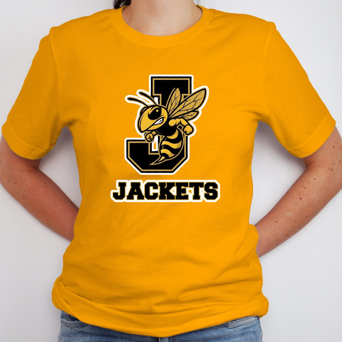 A person wearing a bright yellow short-sleeve t-shirt with a black and yellow 'J' and bee graphic, with the word 'JACKETS' printed below in black with a white outline. The person is also wearing blue jeans against a plain light gray background.