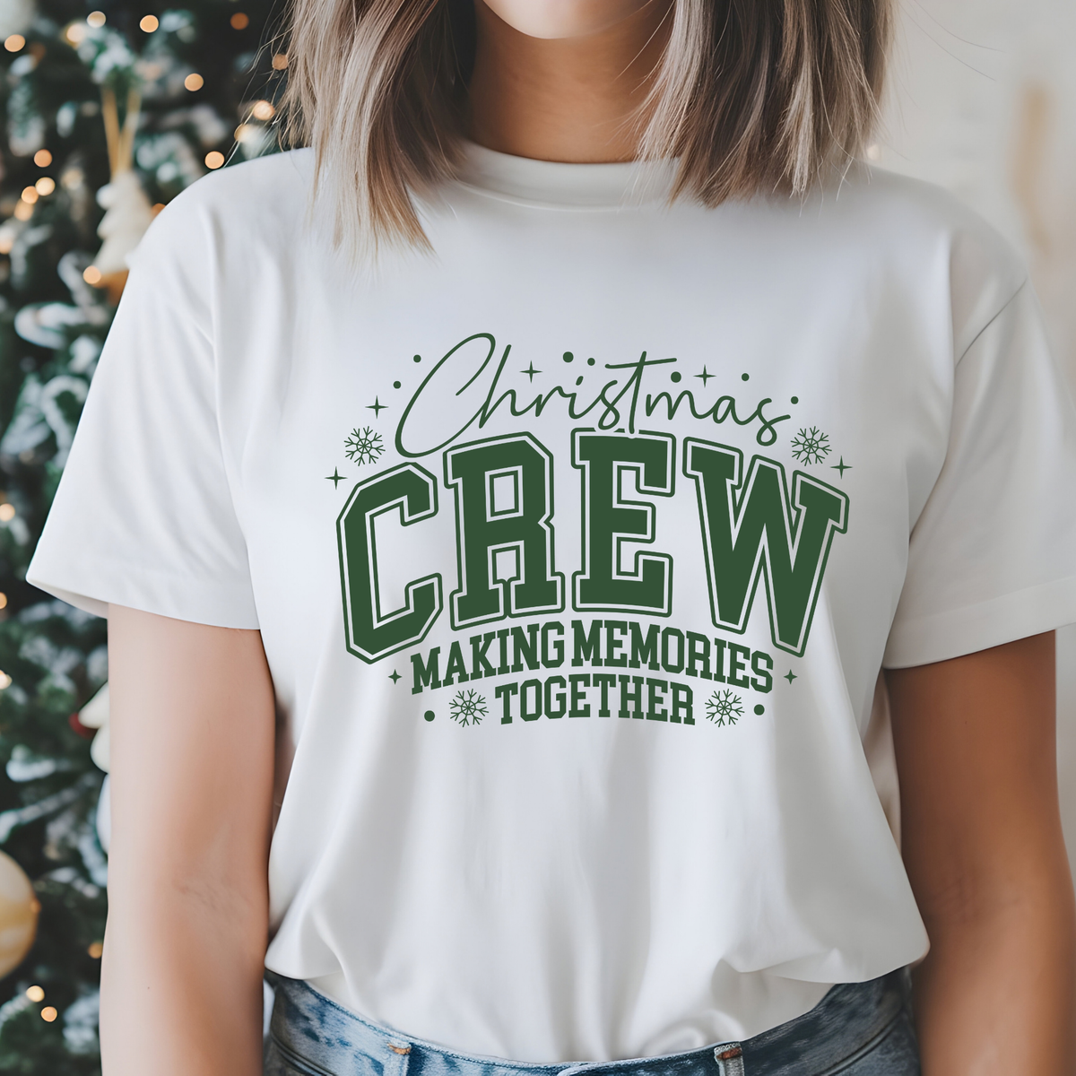 A woman wearing a white crew-neck t-shirt with a dark green graphic that reads 'Christmas CREW MAKING MEMORIES TOGETHER' with snowflake accents, set against a softly blurred holiday background.