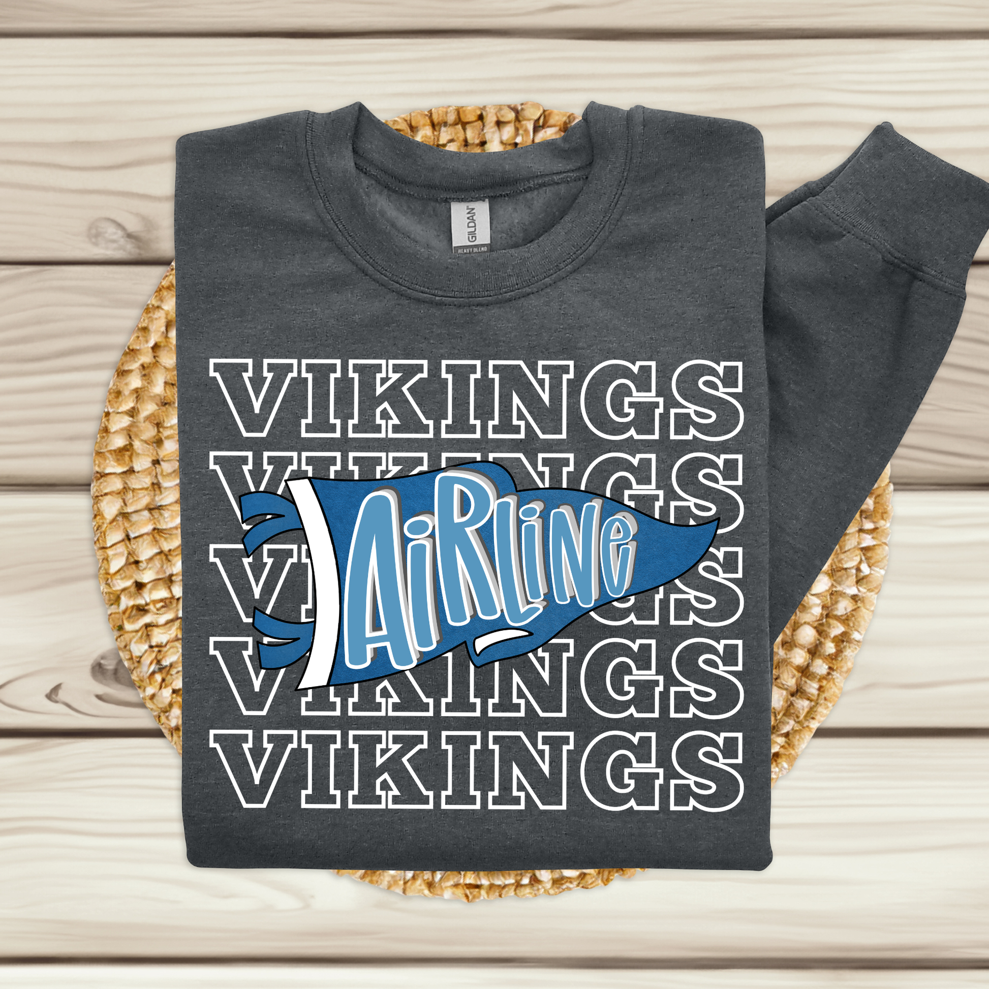 A dark gray long-sleeve t-shirt with a 'Vikings Airline' graphic, laid flat on a woven round mat over a light wood plank background.