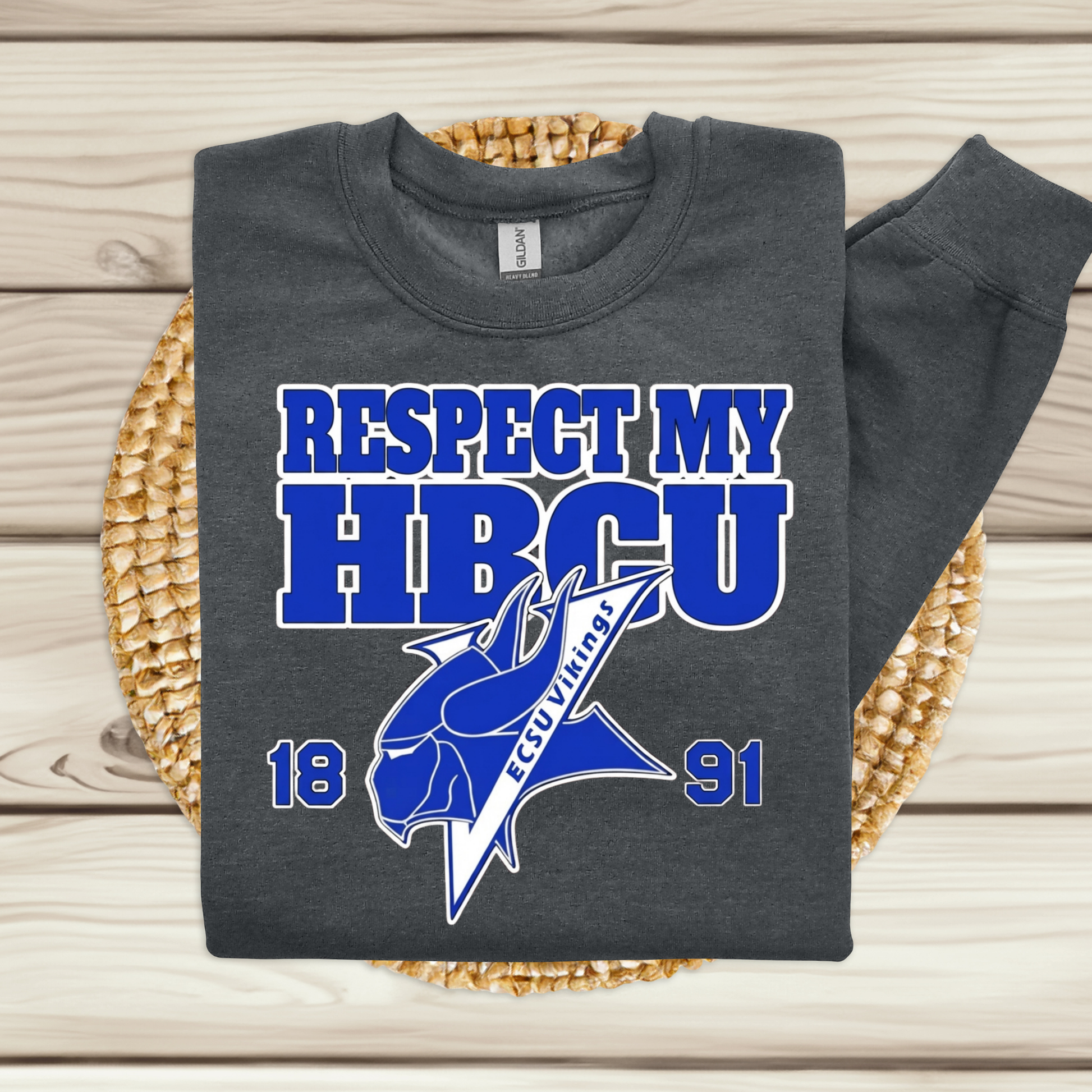 A dark charcoal gray sweatshirt with a blue and white graphic reading 'RESPECT MY HBCU' and '1891' with a fish logo, laid flat on a woven round mat over light wood planks.