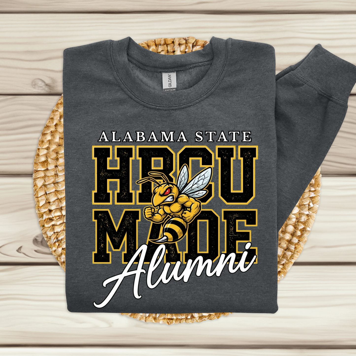 A folded dark gray crewneck sweatshirt with a graphic print reading 'ALABAMA STATE HBCU MADE Alumni' and a bee mascot, displayed on a woven round mat over a light wood plank background.