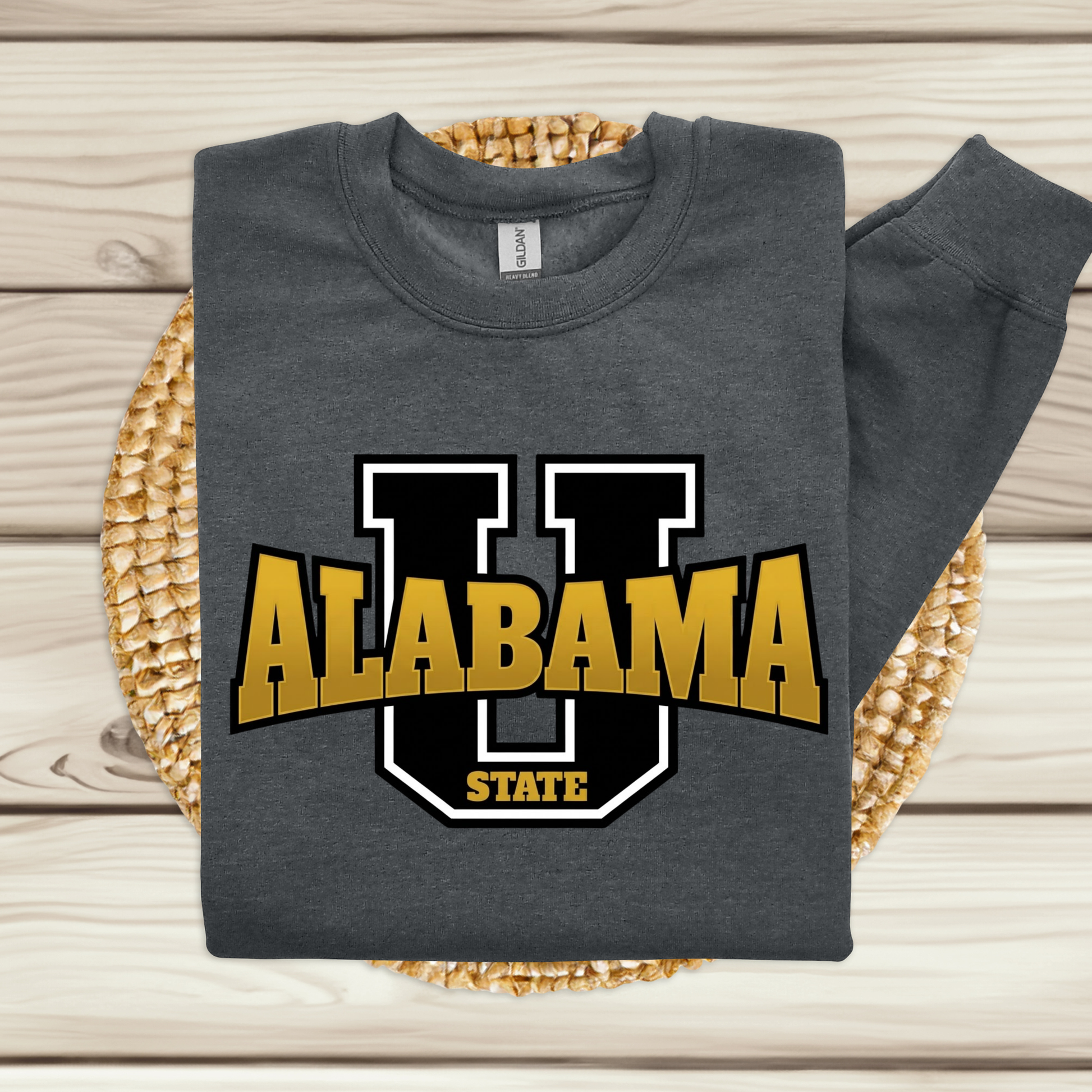 A dark gray long-sleeve sweatshirt laid flat on a woven round mat over a light wood plank surface. The sweatshirt features a large front graphic with a black and white outlined letter U, the word ALABAMA in yellow with black outlining, and the word STATE below in yellow.