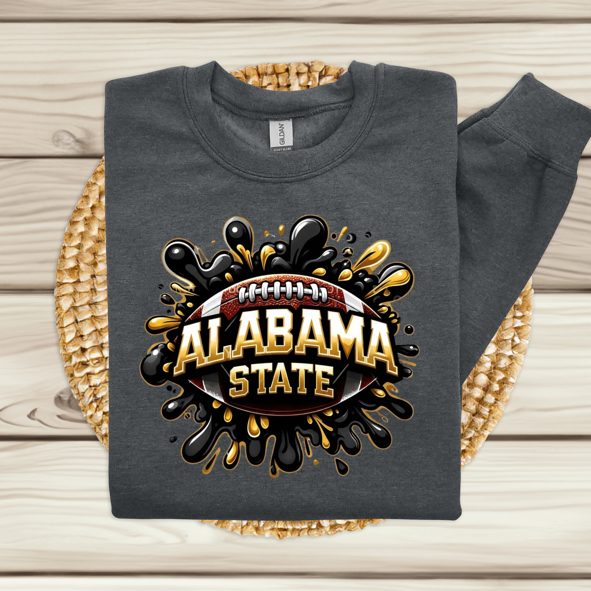 A dark gray sweatshirt laid flat on a woven round mat over a light wood plank surface. The sweatshirt has a large front graphic featuring a football and the words 'ALABAMA STATE' in gold and black paint-splatter style.