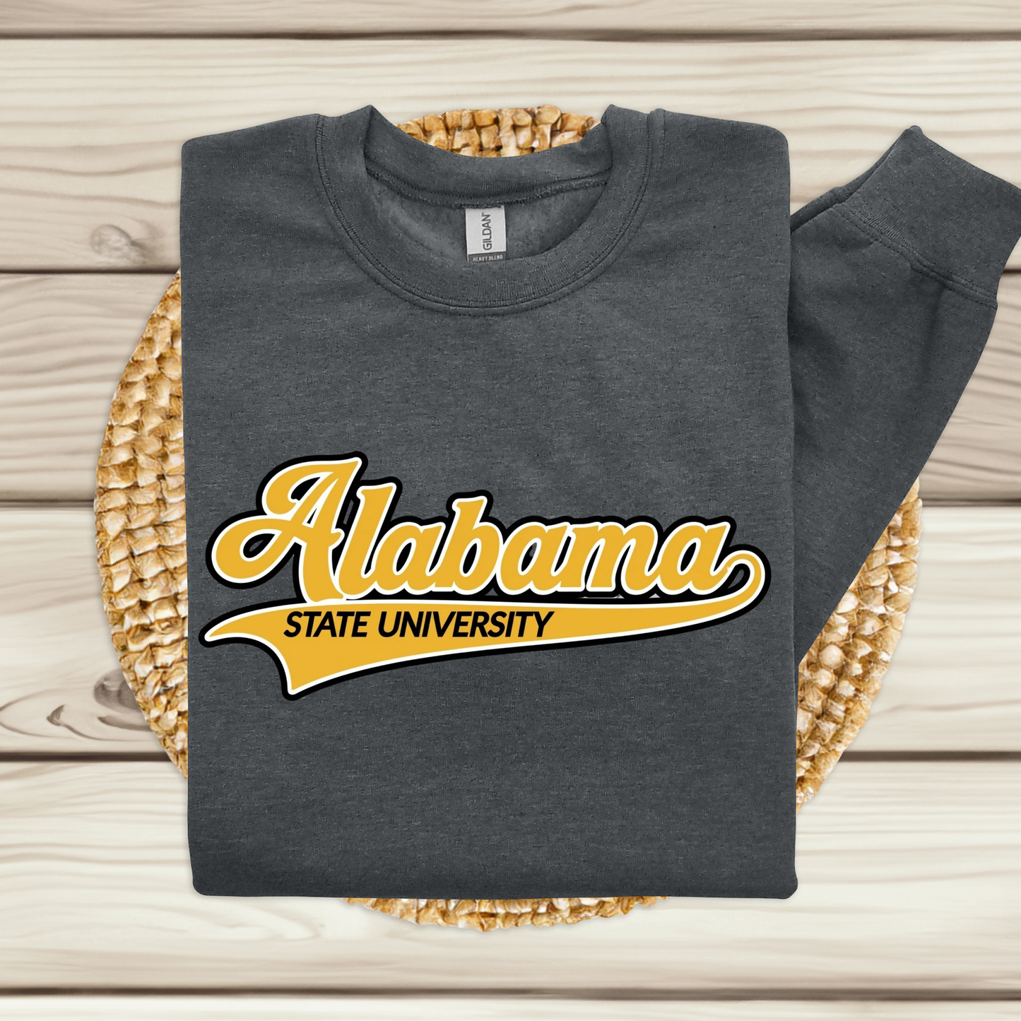 A dark gray long-sleeve crewneck sweatshirt with a yellow and white 'Alabama State University' graphic on the front, styled flat on a woven placemat over a light wood plank background.