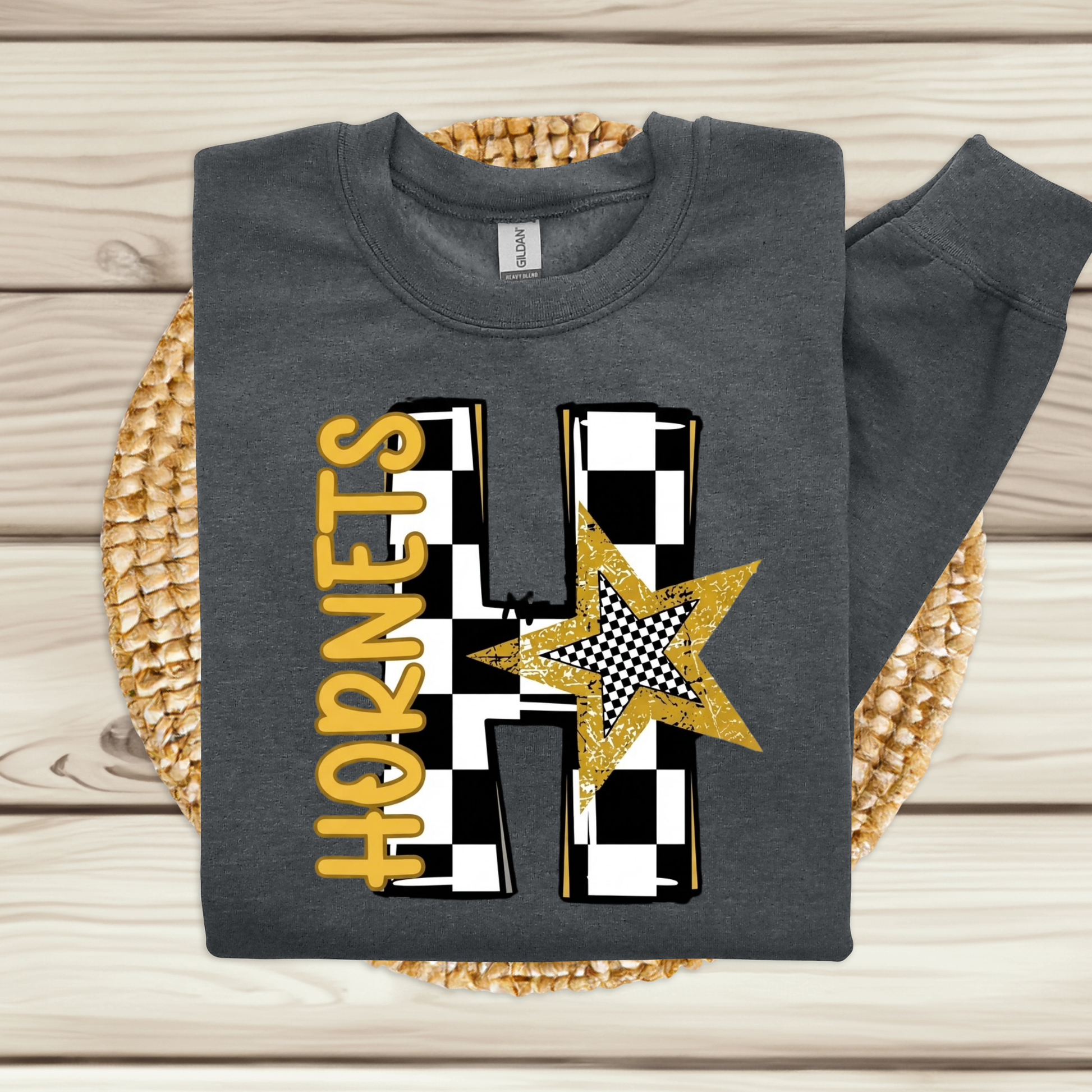 A dark gray sweatshirt laid flat on a woven round mat over a light wood plank surface. The sweatshirt has a large front graphic featuring the word 'HORNETS' in yellow, a black-and-white checkered letter H, and a gold glitter star.