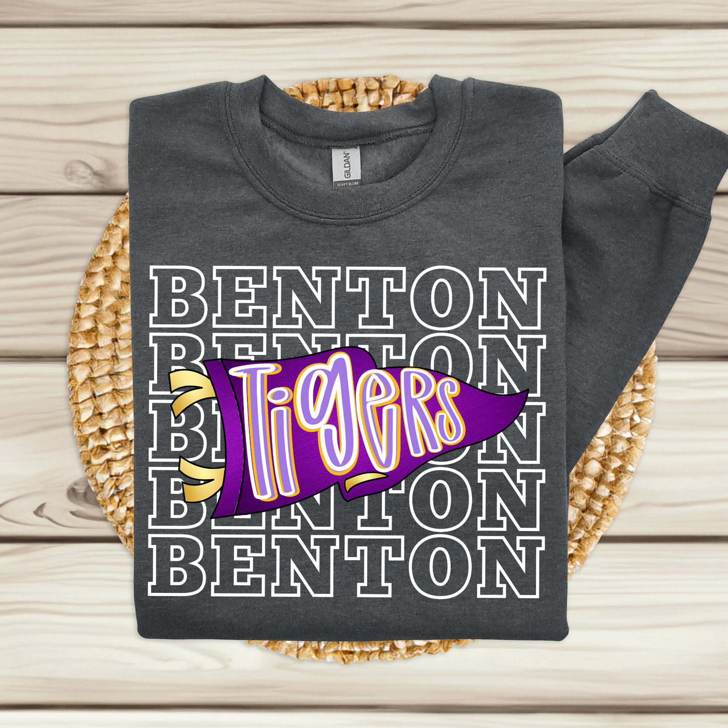 A dark charcoal gray sweatshirt laid flat on a woven round mat over a light wood plank surface. The sweatshirt features a graphic design with the word 'BENTON' repeated in white outlined letters, overlaid with a purple pennant flag containing the word 'Tigers' in yellow and white with a purple outline.