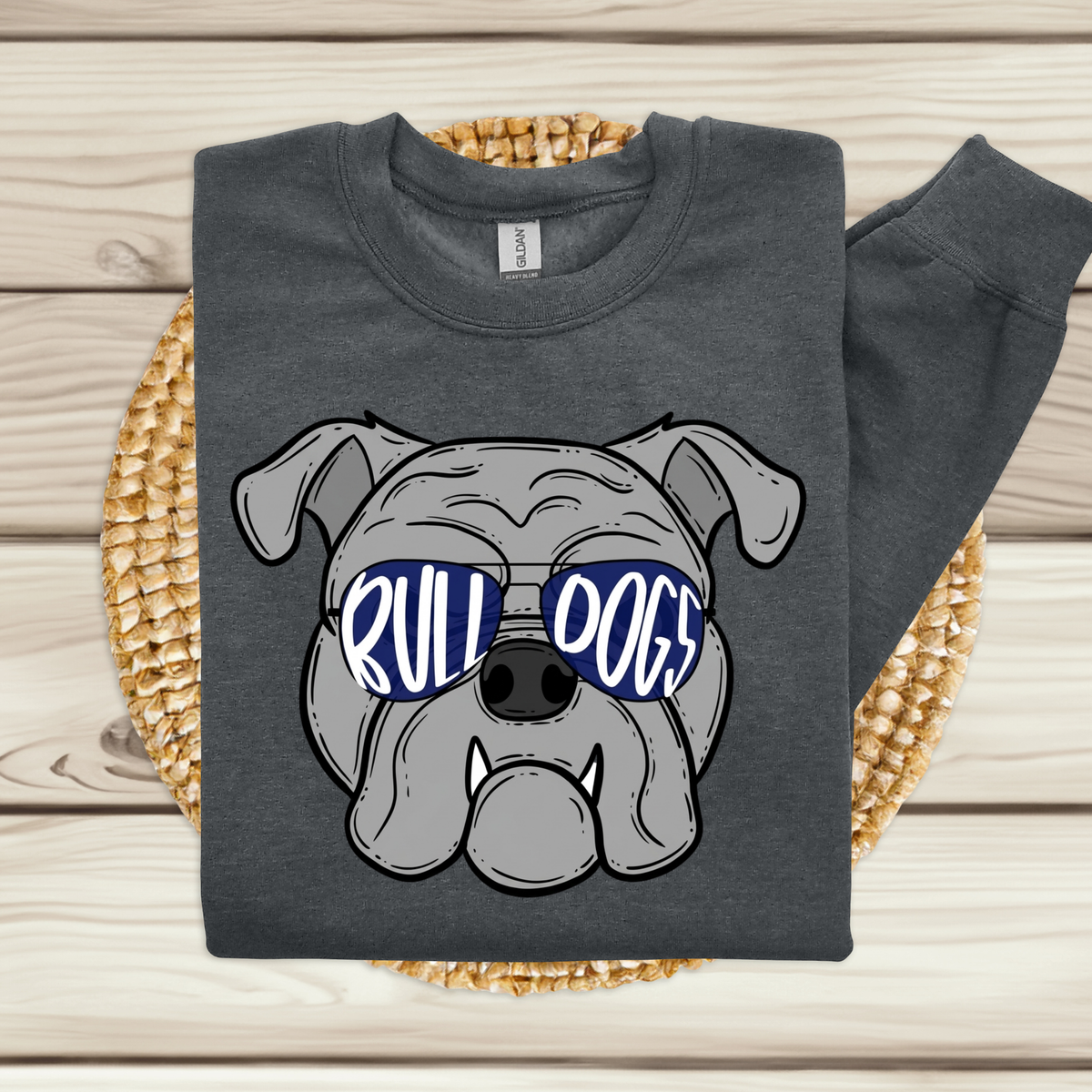 A dark gray sweatshirt laid flat on a woven round mat over a light wood plank surface. The sweatshirt has a large graphic of a bulldog wearing blue sunglasses with the words 'RULL DOGS' in white.