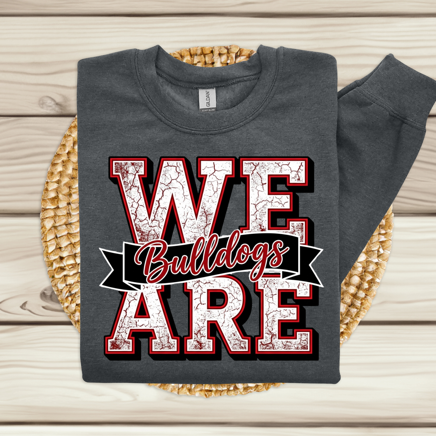 A dark gray sweatshirt laid flat on a woven round mat over a light wood plank surface. The sweatshirt has a large front graphic reading “WE ARE Bulldogs” in distressed white letters outlined in red and black, with a black banner and red script text across the middle.