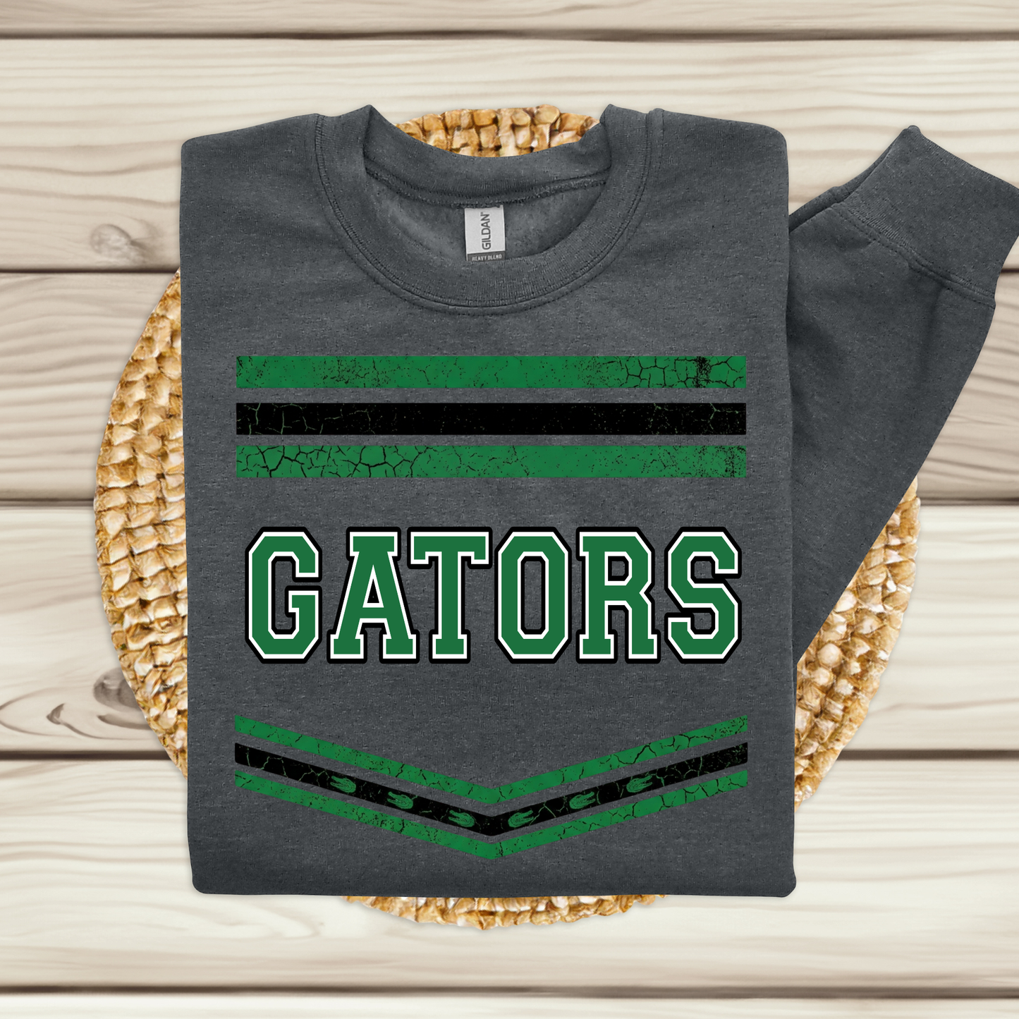 A dark gray sweatshirt laid flat on a woven straw mat over a light wood plank surface. The sweatshirt has green and black horizontal stripes and the word 'GATORS' in large green varsity-style letters outlined in white.
