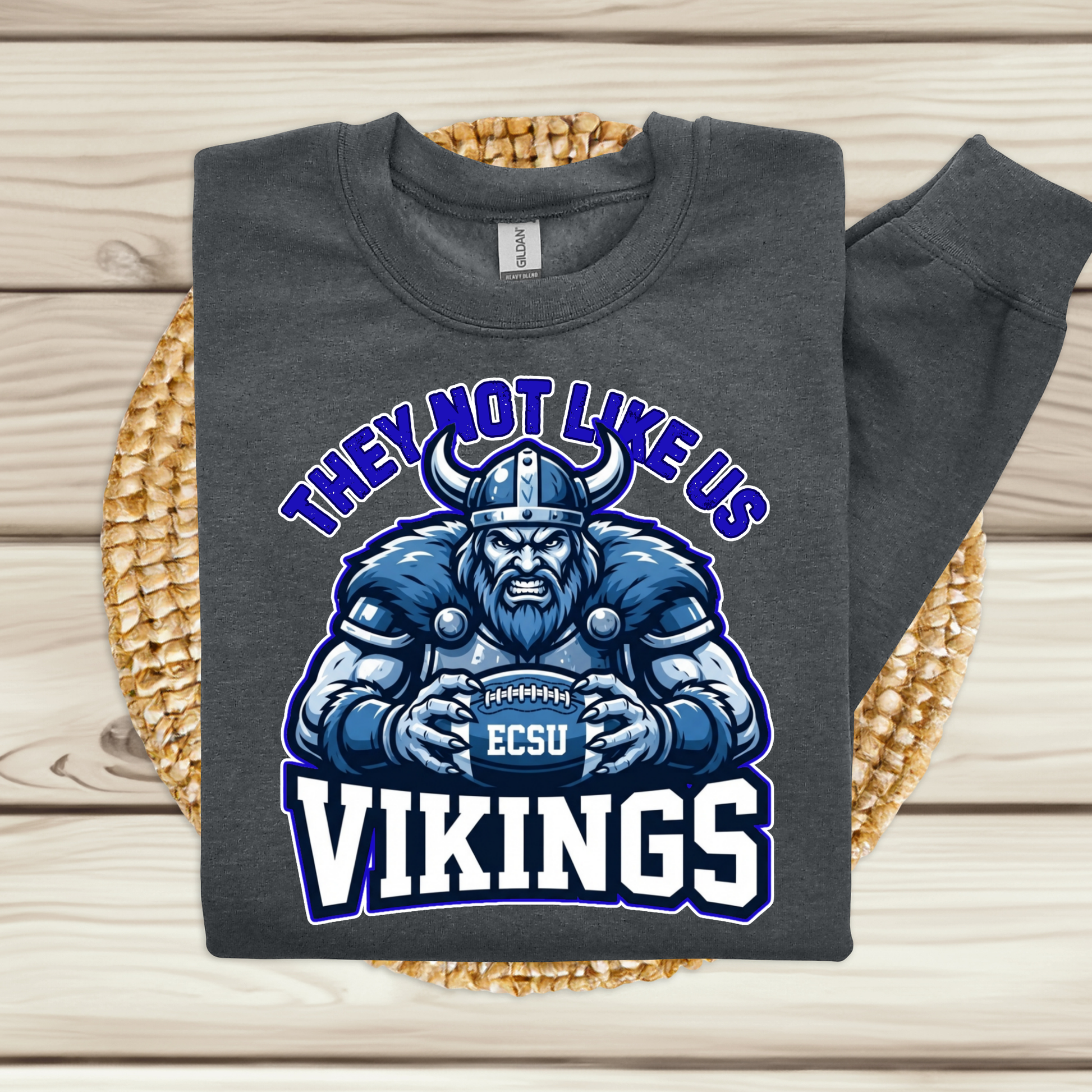 A dark gray sweatshirt laid flat on a woven round mat over a light wood plank surface. The sweatshirt has a large graphic of a blue and white Viking holding a football with the text 'THEY NOT LIKE US VIKINGS' and 'ECSU' on the ball.