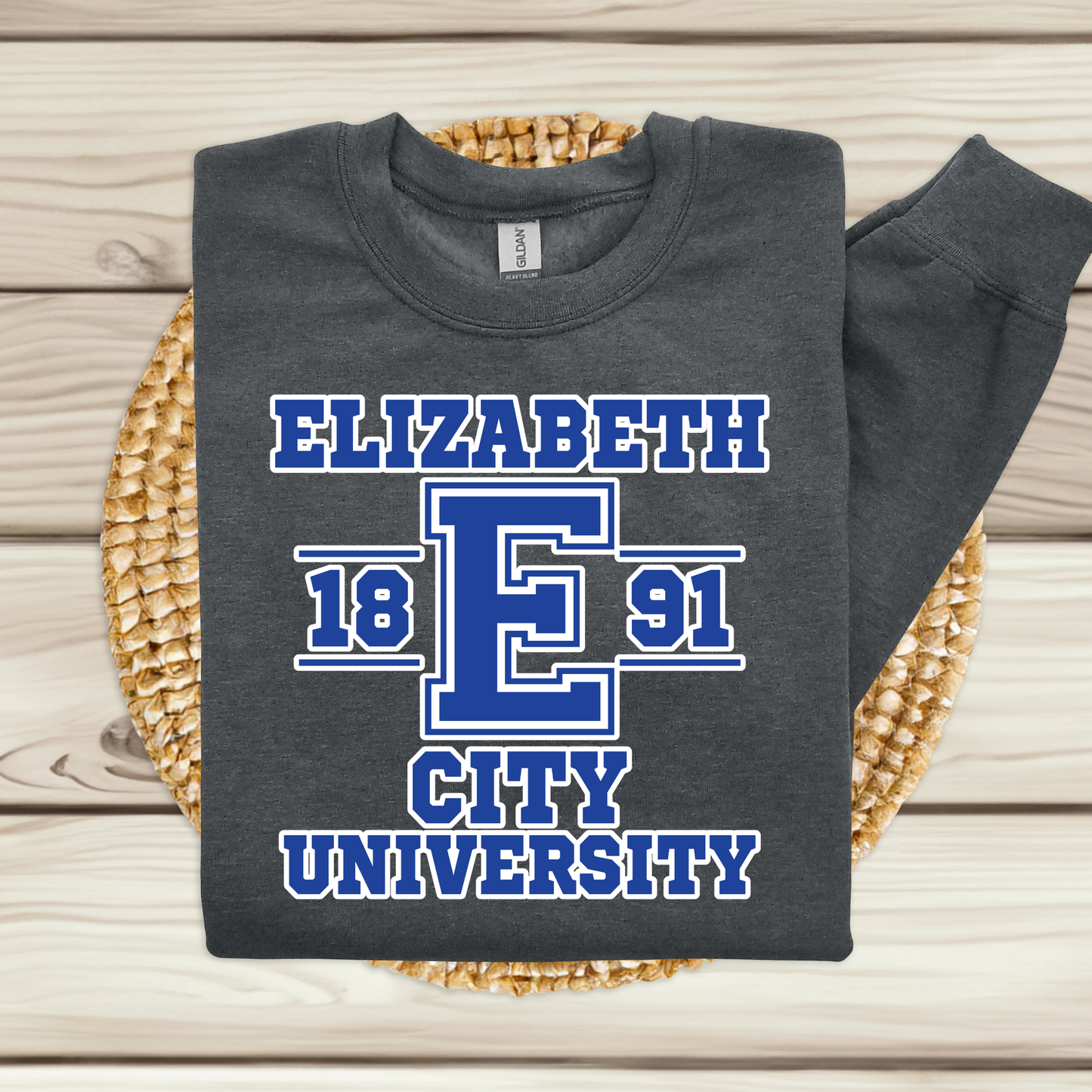A dark charcoal gray sweatshirt with a blue and white graphic reading 'ELIZABETH 18 E 91 CITY UNIVERSITY,' folded and displayed on a woven round mat over a light wood plank background.