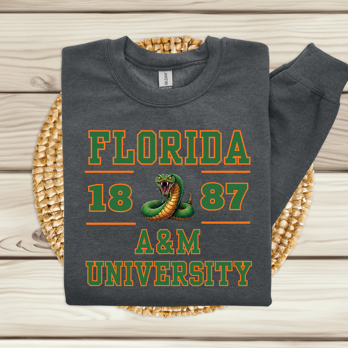 A folded dark gray sweatshirt with a long sleeve, featuring a green and orange graphic that reads 'FLORIDA 1887 A&M UNIVERSITY' with a coiled snake in the center, displayed on a woven round mat over a light wood plank background.
