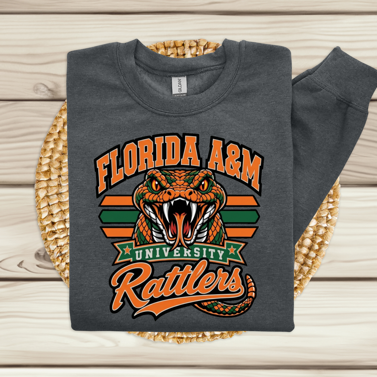 A folded dark gray long-sleeve sweatshirt with a Florida A&M University Rattlers graphic, displayed on a woven round mat over a light wood plank background.